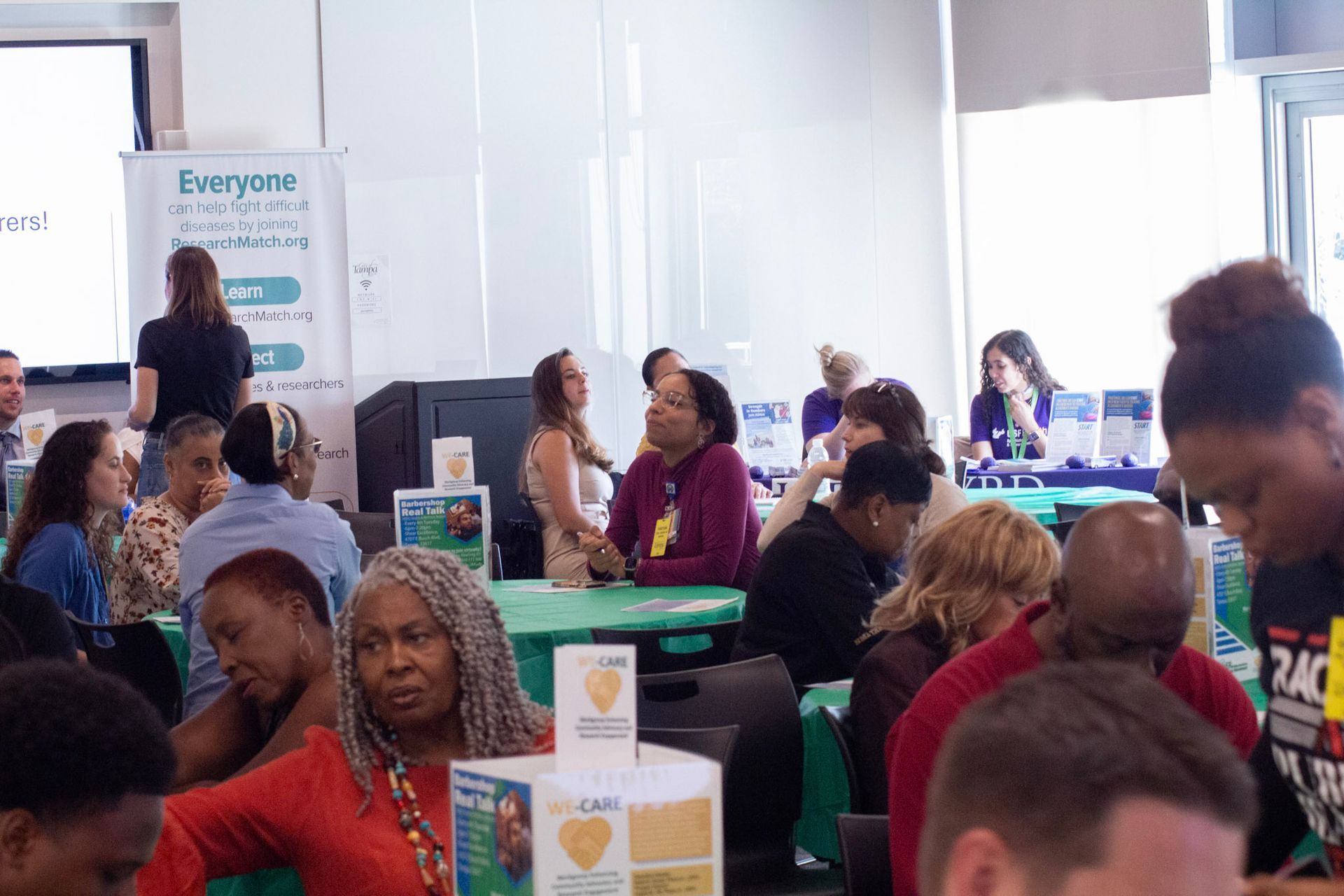 People gathered at tables in a brightly lit room; one person speaks near a sign.
