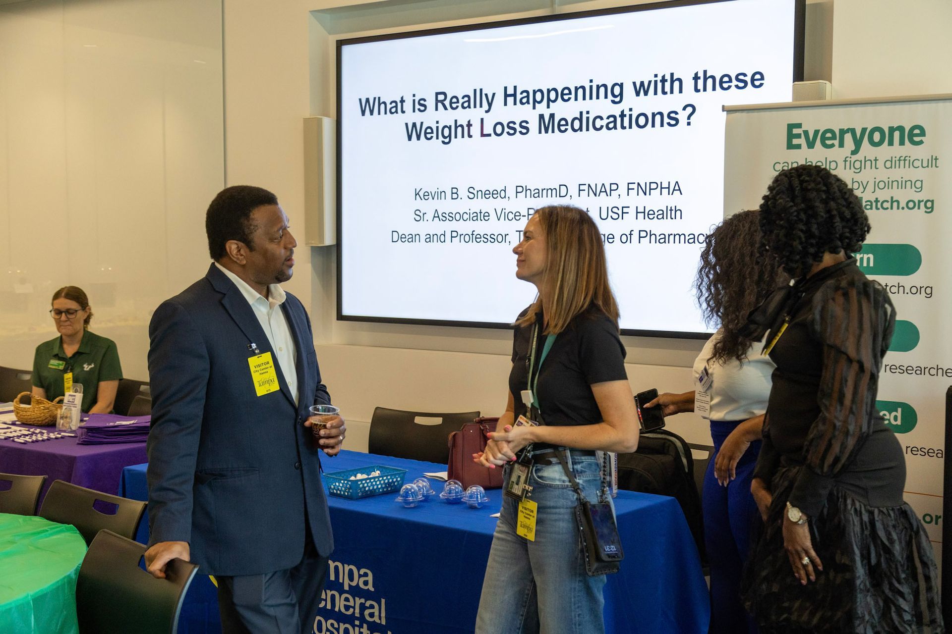 People at an event about weight-loss medications. A man speaks, two women listen, a presentation screen is behind them.