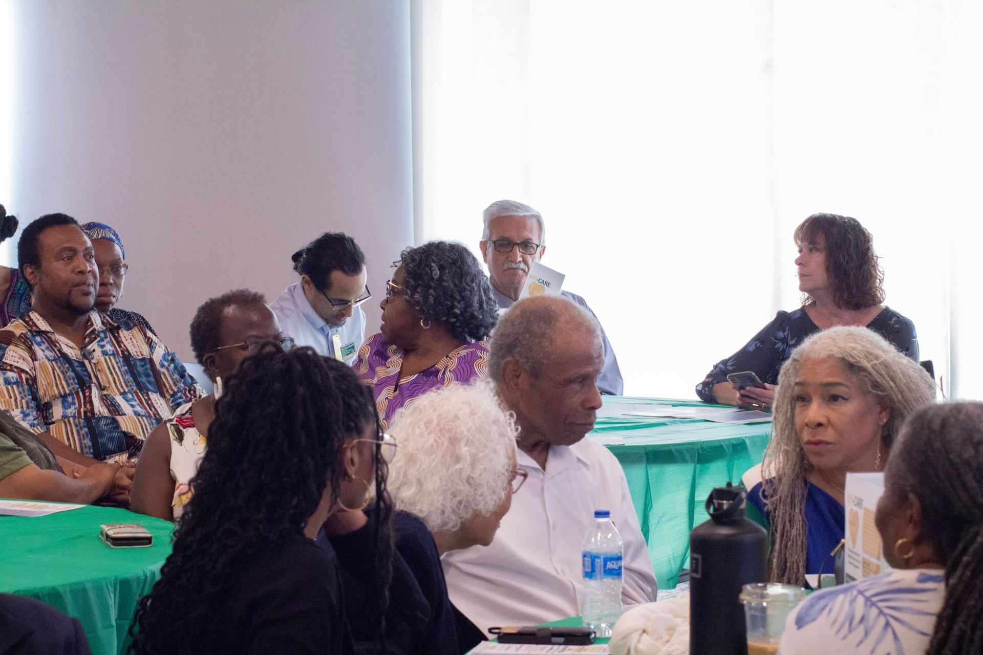 People seated at tables, some talking, in a brightly lit room with a window, possibly a meeting.