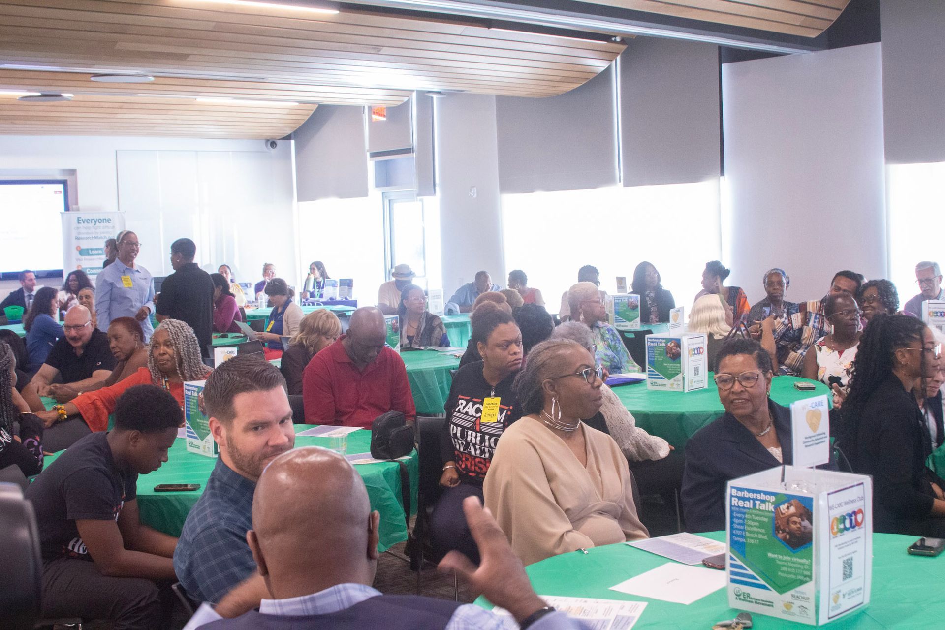 People seated at tables in a conference room, listening to a speaker. Green tablecloths and white walls.