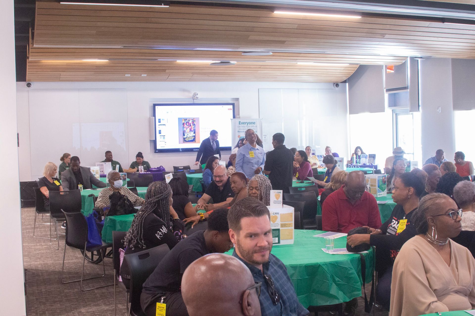 People seated at round green tables, attending a conference in a well-lit room with a large screen.