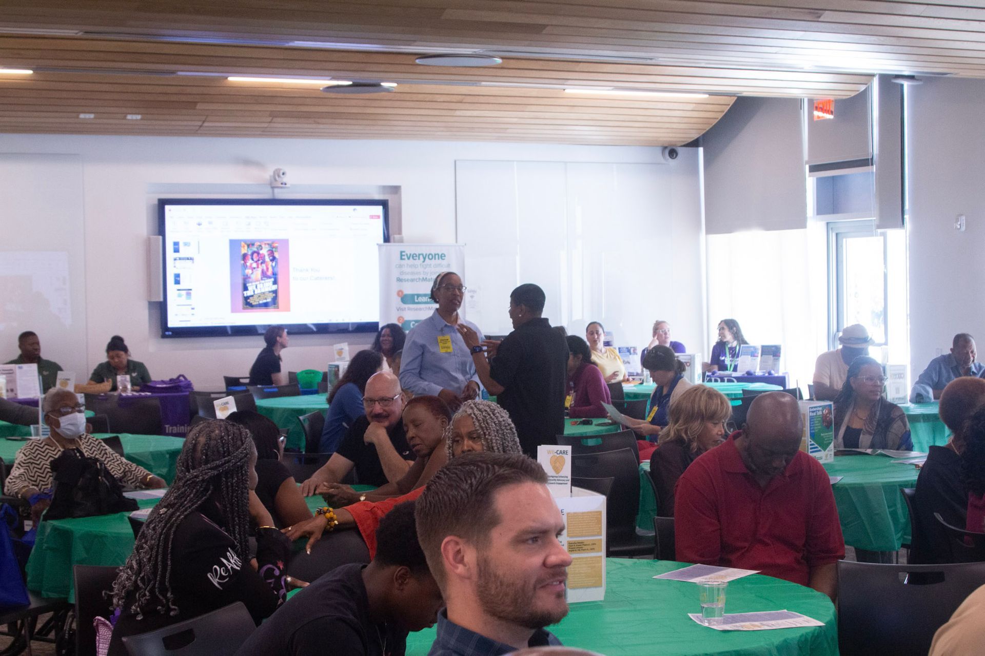 Attendees at a conference seated at tables with green tablecloths; presenter standing with microphone.