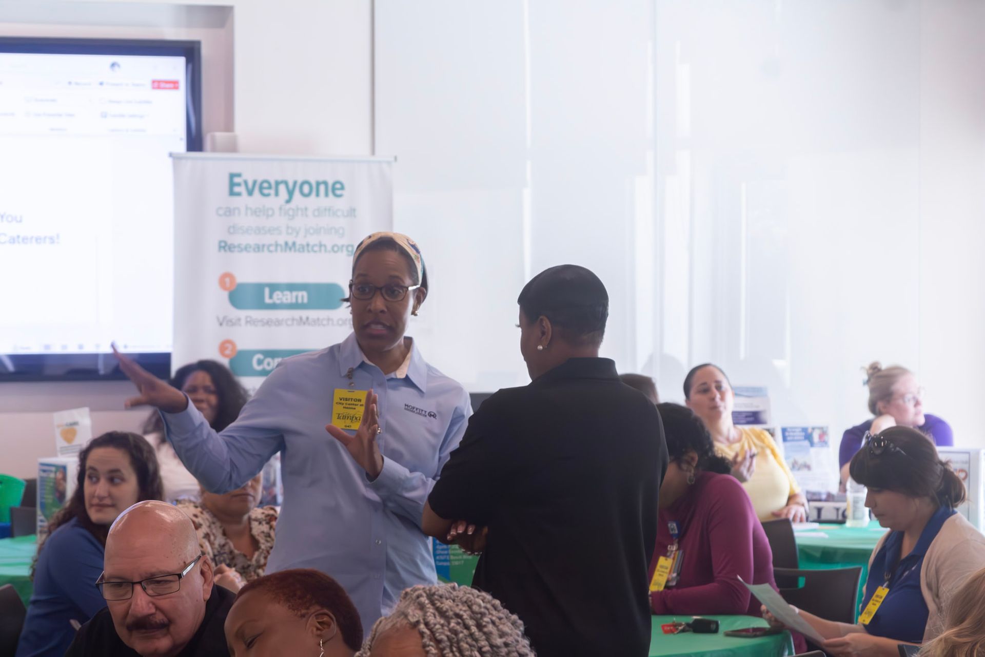 Woman gestures with a sticky note, speaking to another person in a room with a presentation banner.
