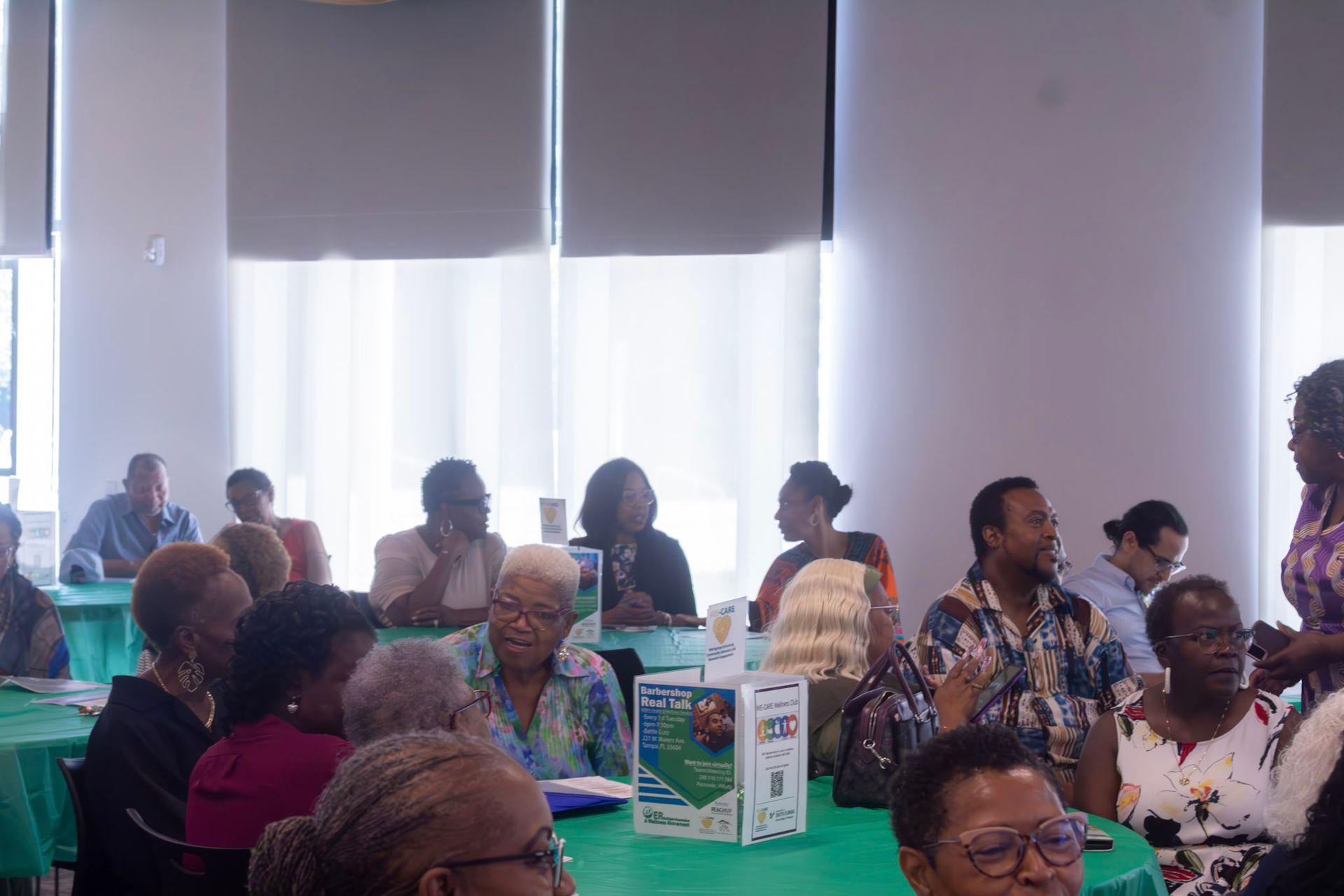 People seated at round tables in a brightly lit room, some looking at a speaker, others looking at each other.