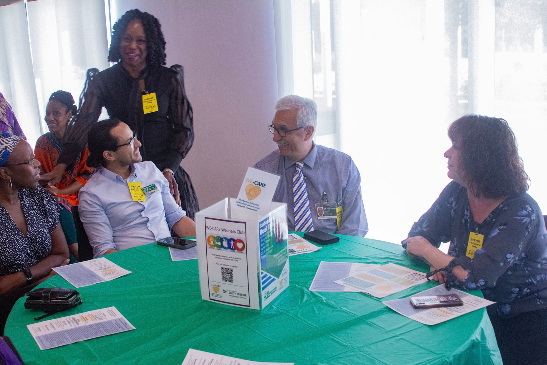 People at a round table with a green cover, discussing; a ballot box sits in the center.