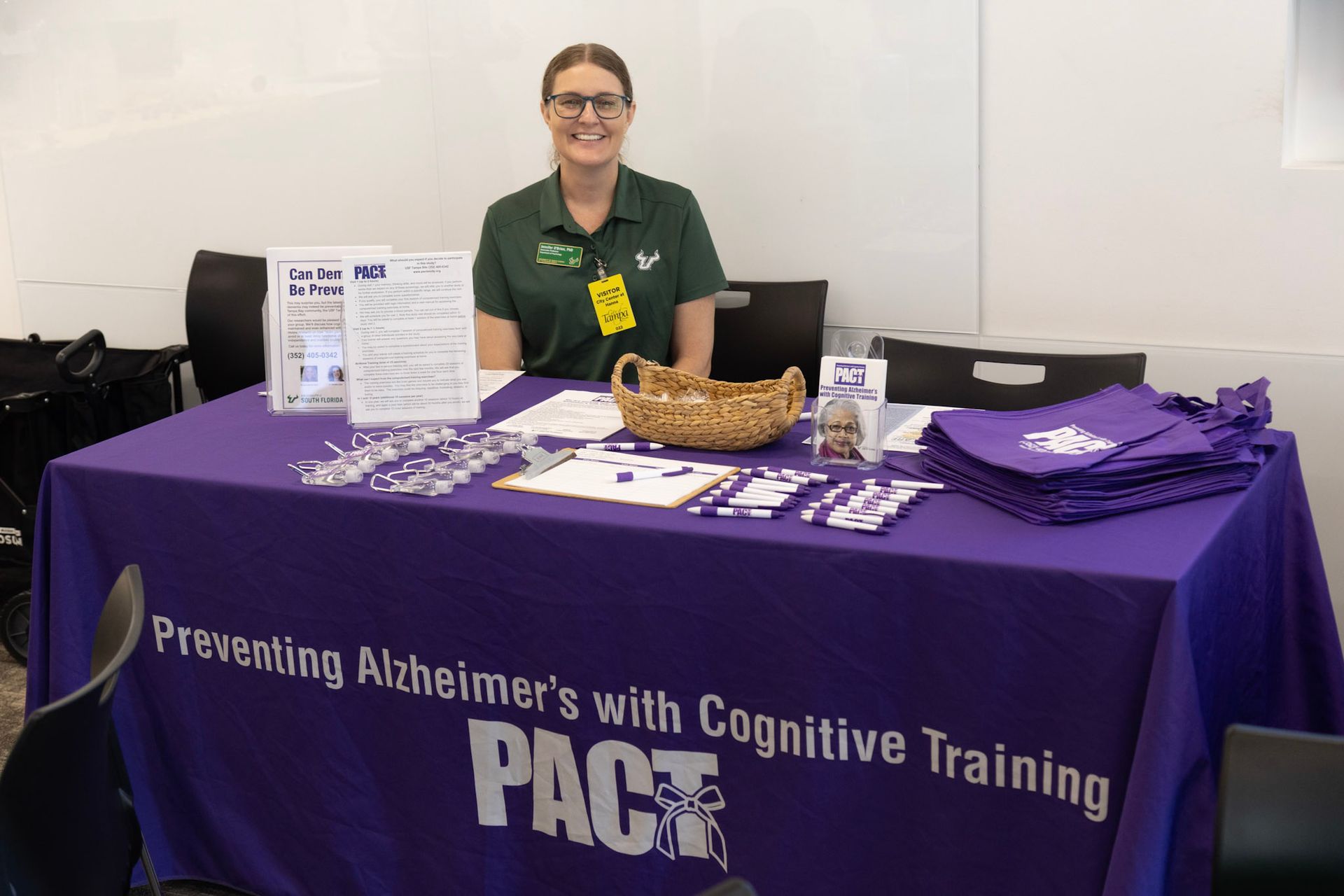 Woman at PACT table, smiling. Table has purple cover, materials about preventing Alzheimer's, and a basket.