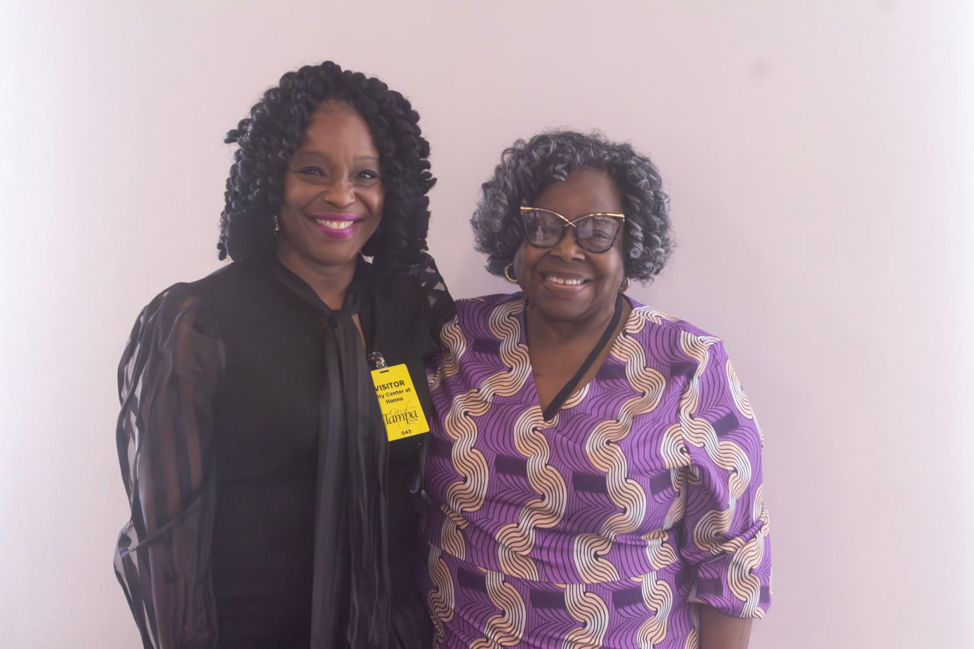 Two women pose, smiling. One in black, one in purple patterned top, standing against a pink wall.