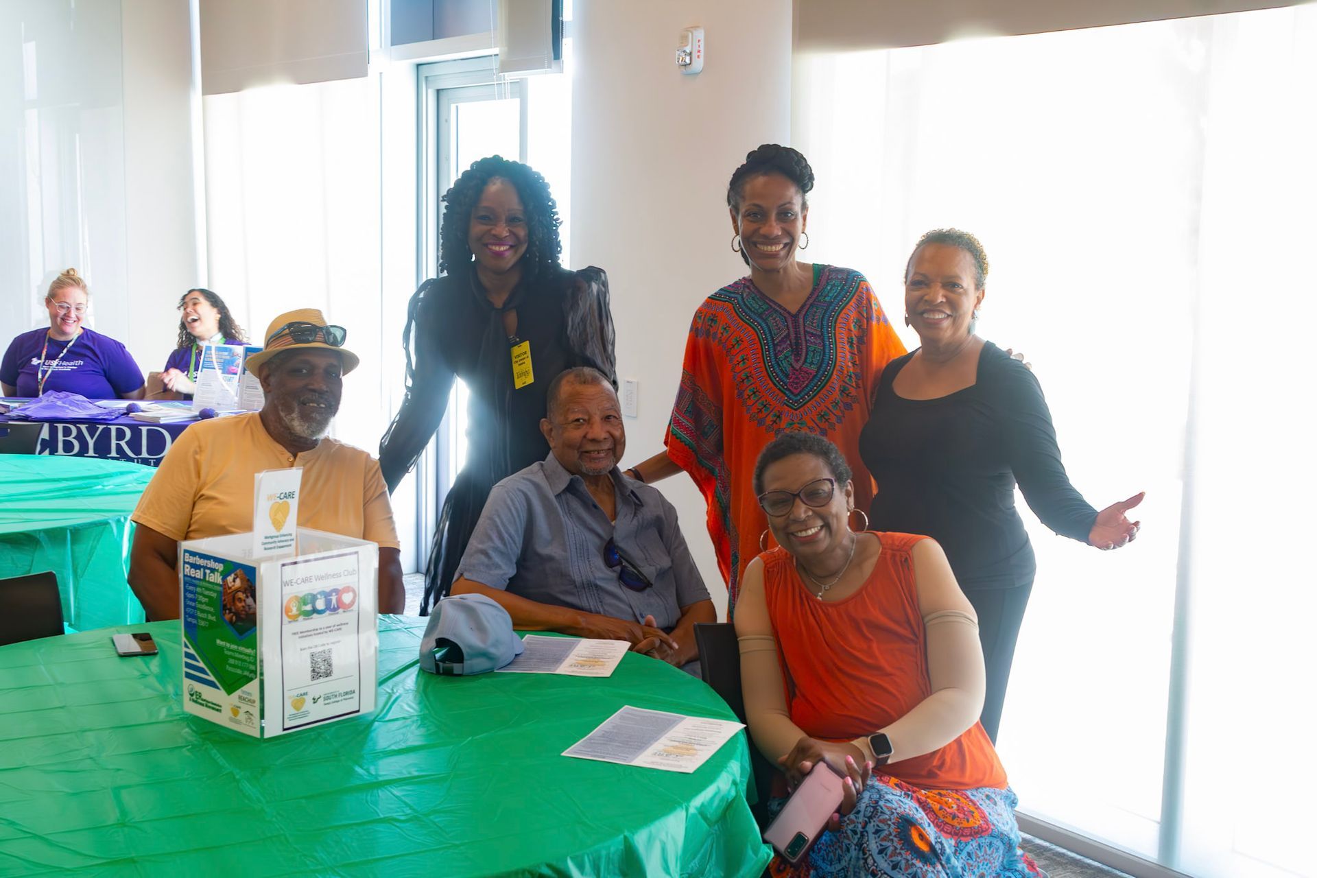 Group of people smiling, posing around green tables. Sunlight streams through a window.