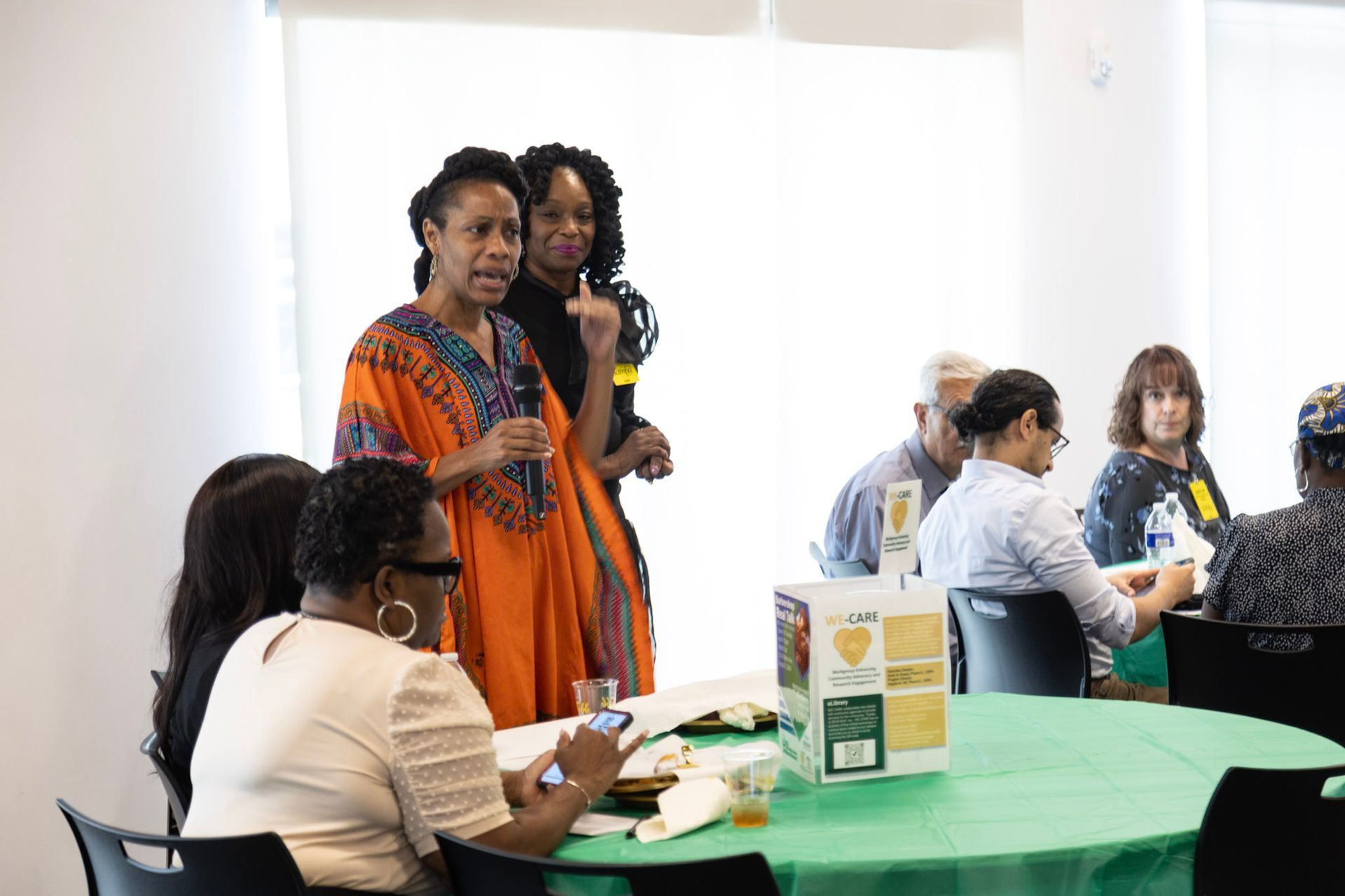 Two women speaking at a table with a group, woman in orange dress speaks into a microphone.