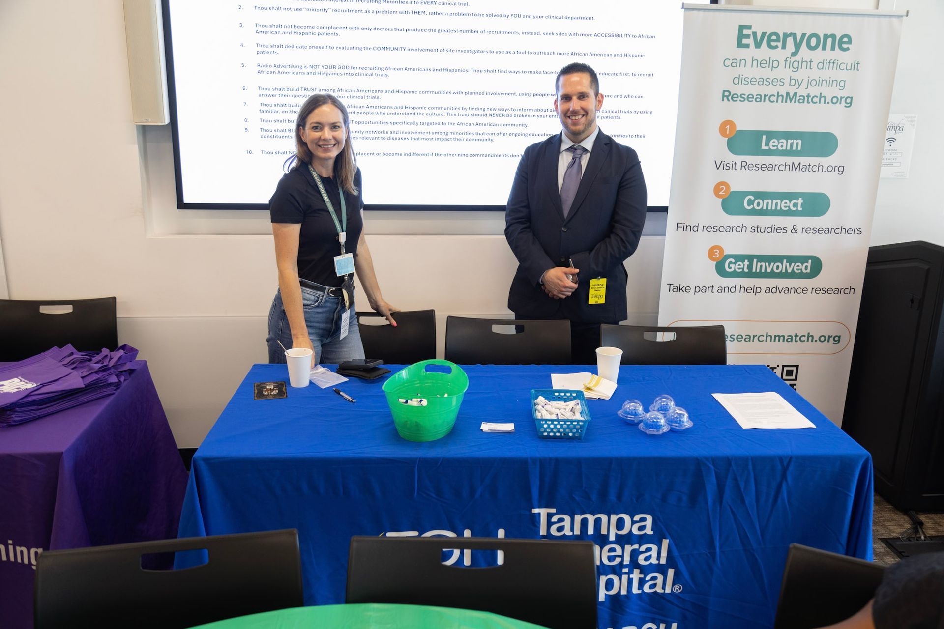 Two people stand behind a Tampa General Hospital information table, smiling. A banner promotes community involvement.
