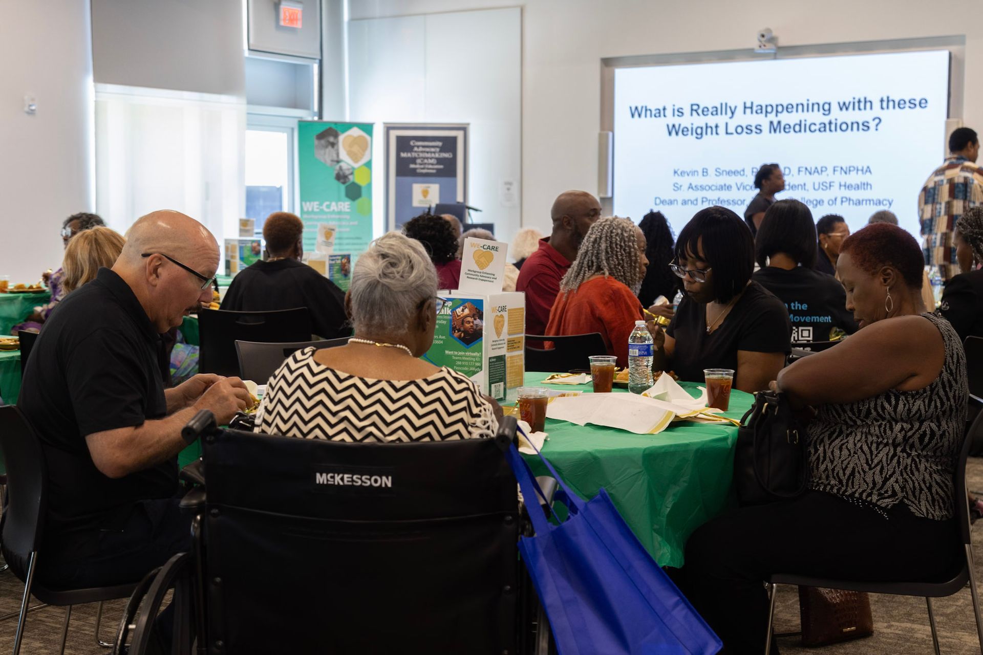 Audience at a seminar, seated at tables with green tablecloths. Presentation title: 