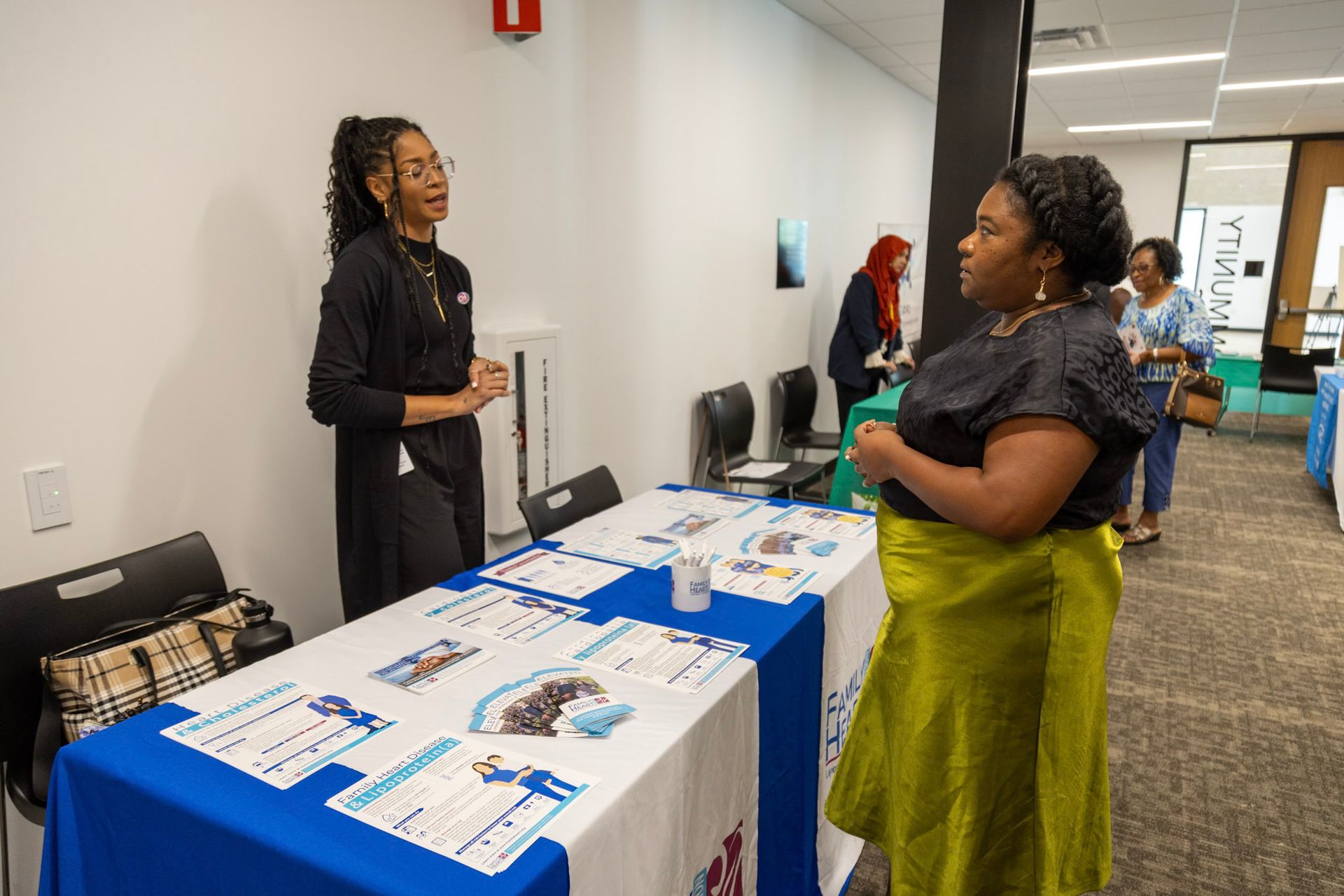 A person at a table talks to another at an event. Flyers on the table. White walls, bright room.