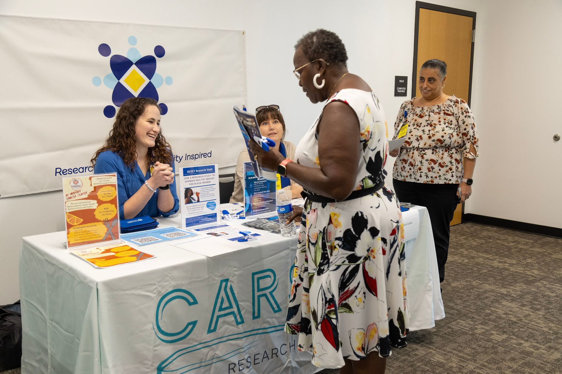 Woman at a booth looking at brochures; another woman stands nearby. Bright, inviting colors, indoors.