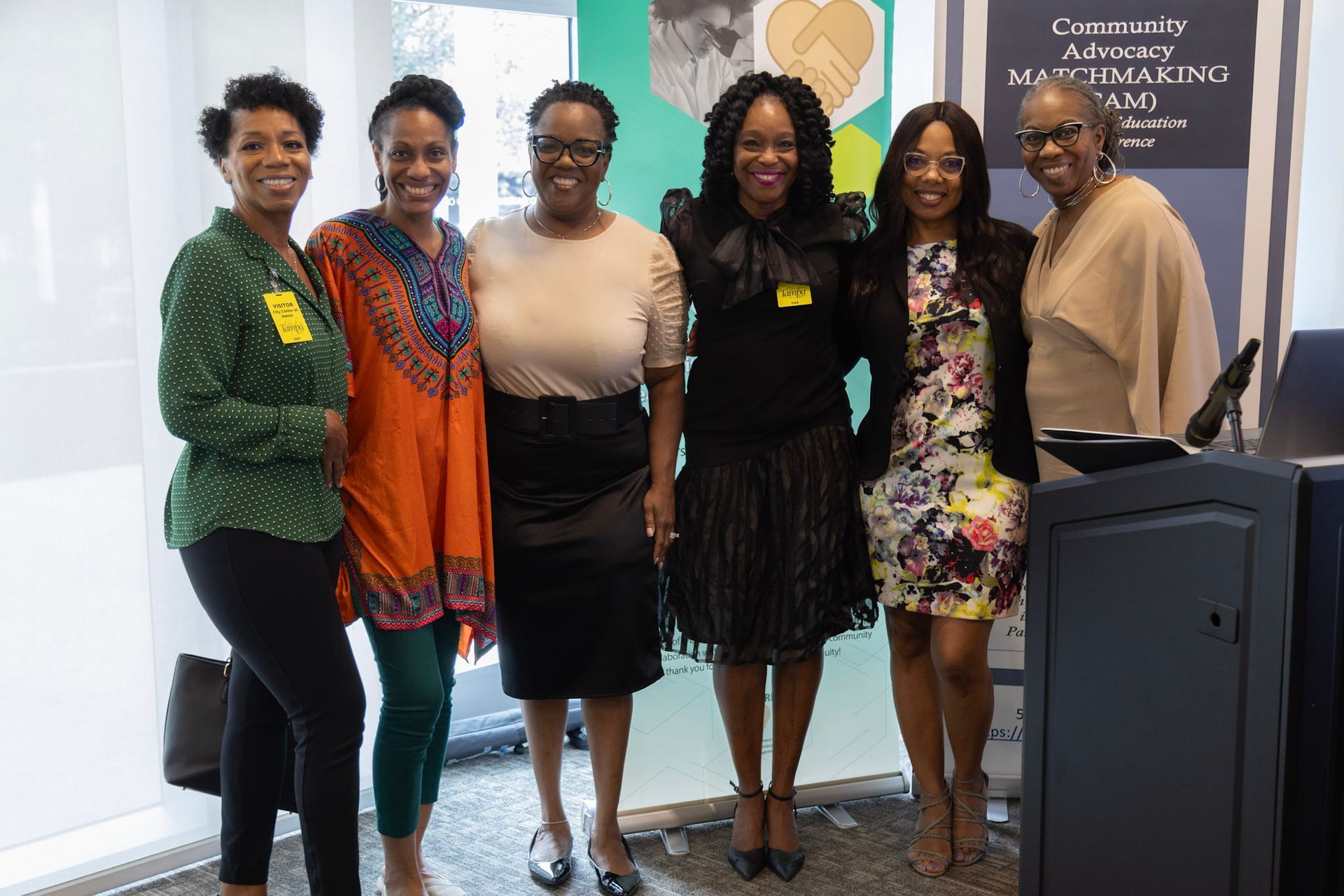 Six women standing together, smiling, at an event. They are near a podium and a sign.