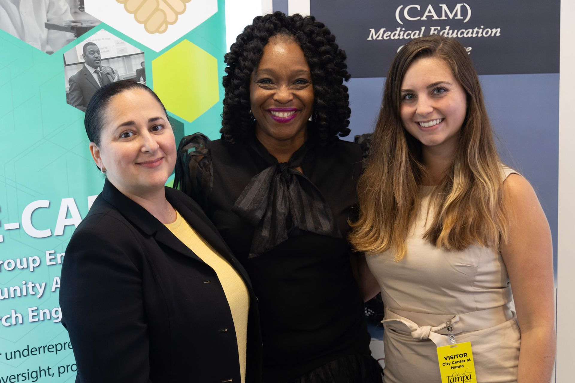 Three women pose together; the central one wears a black dress, smiling.  They stand near a banner.