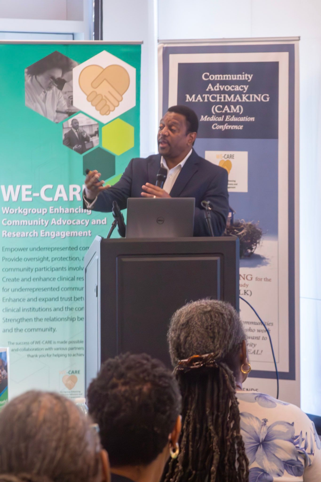 Man speaking at a podium, gesturing. Audience members in foreground. Banners with text and logos on the sides.