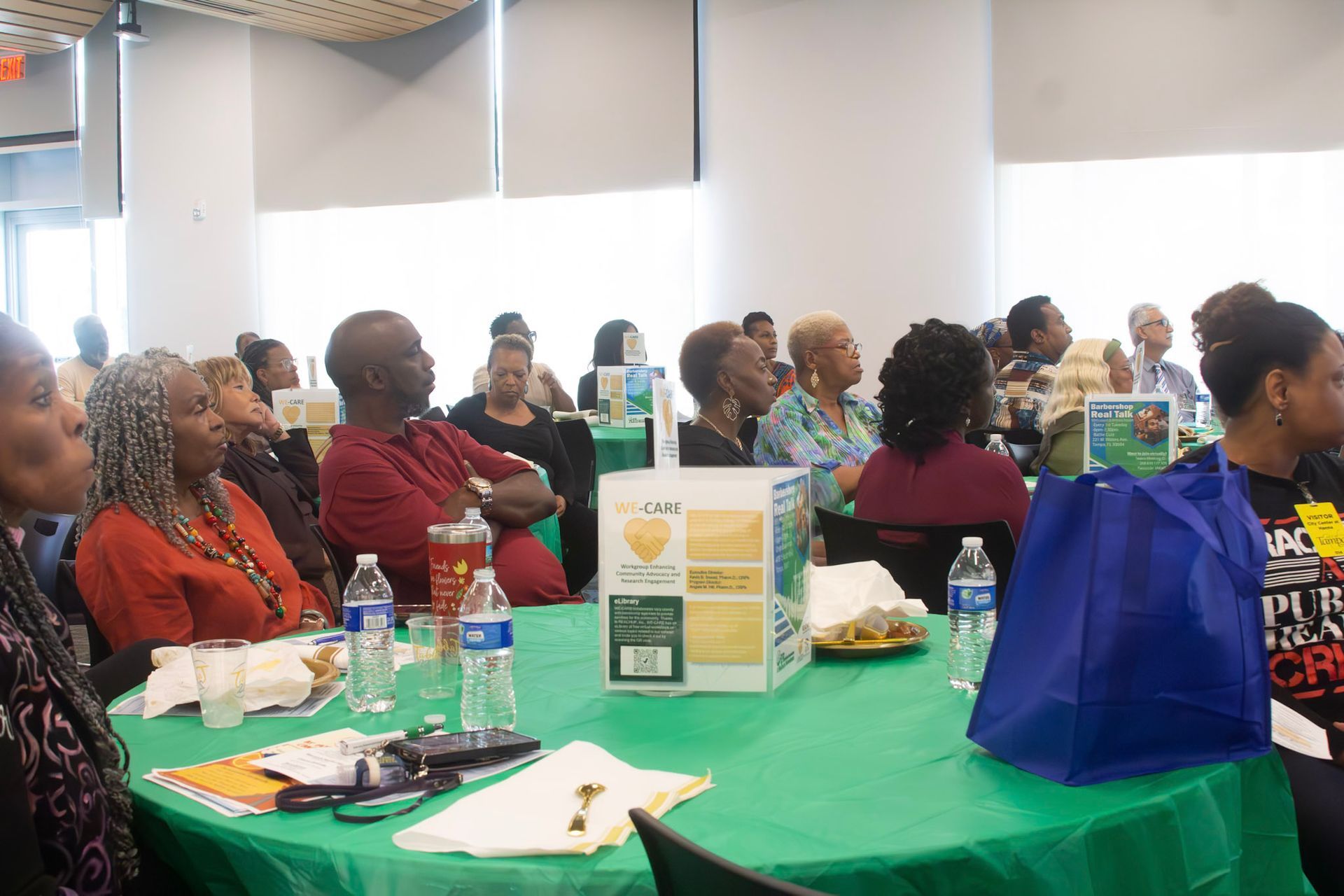 Audience at tables, attending an event. Green tablecloths, water bottles, and a blue tote bag.