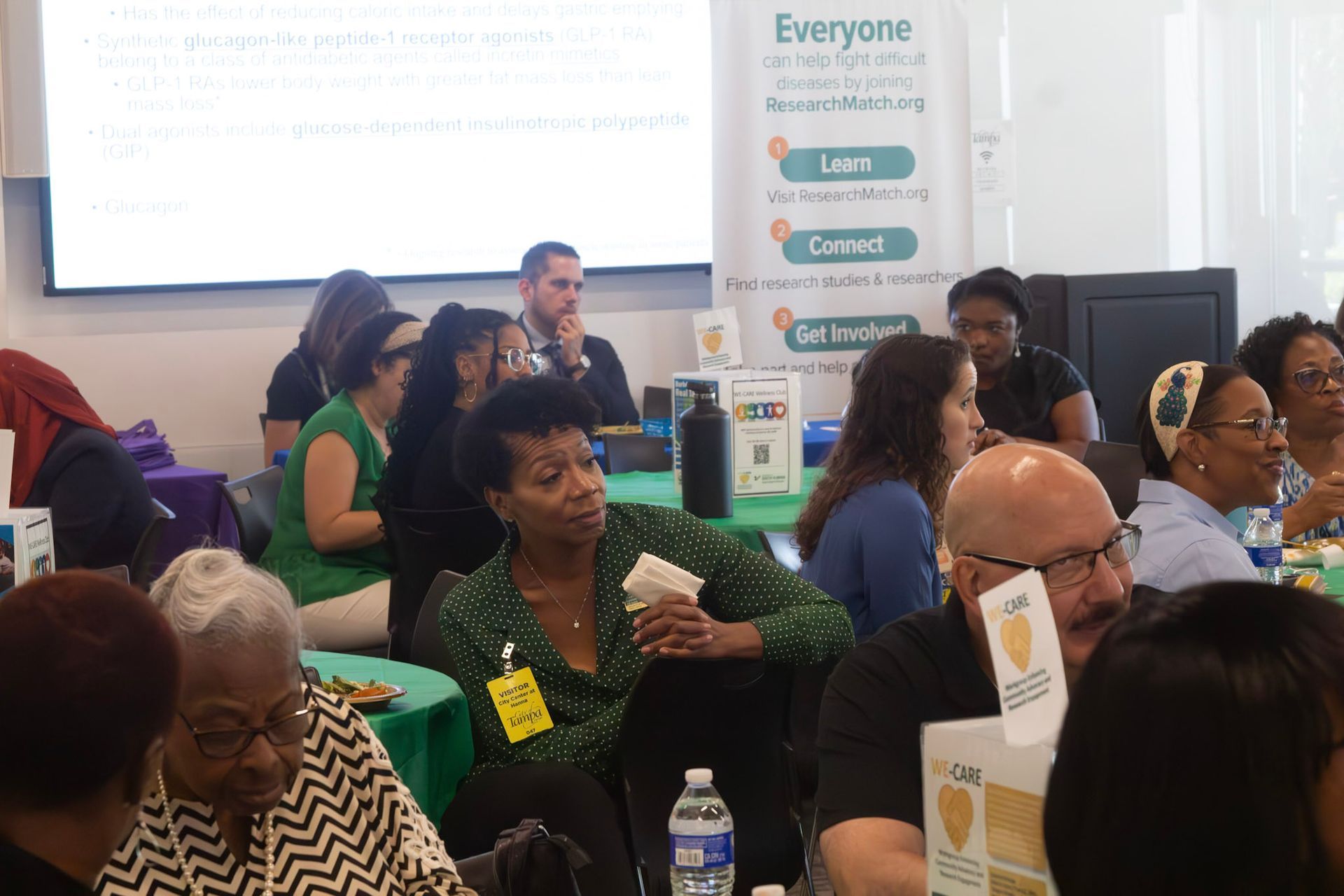 People seated at tables in a conference room. Some are talking, some looking ahead. A banner in the background.