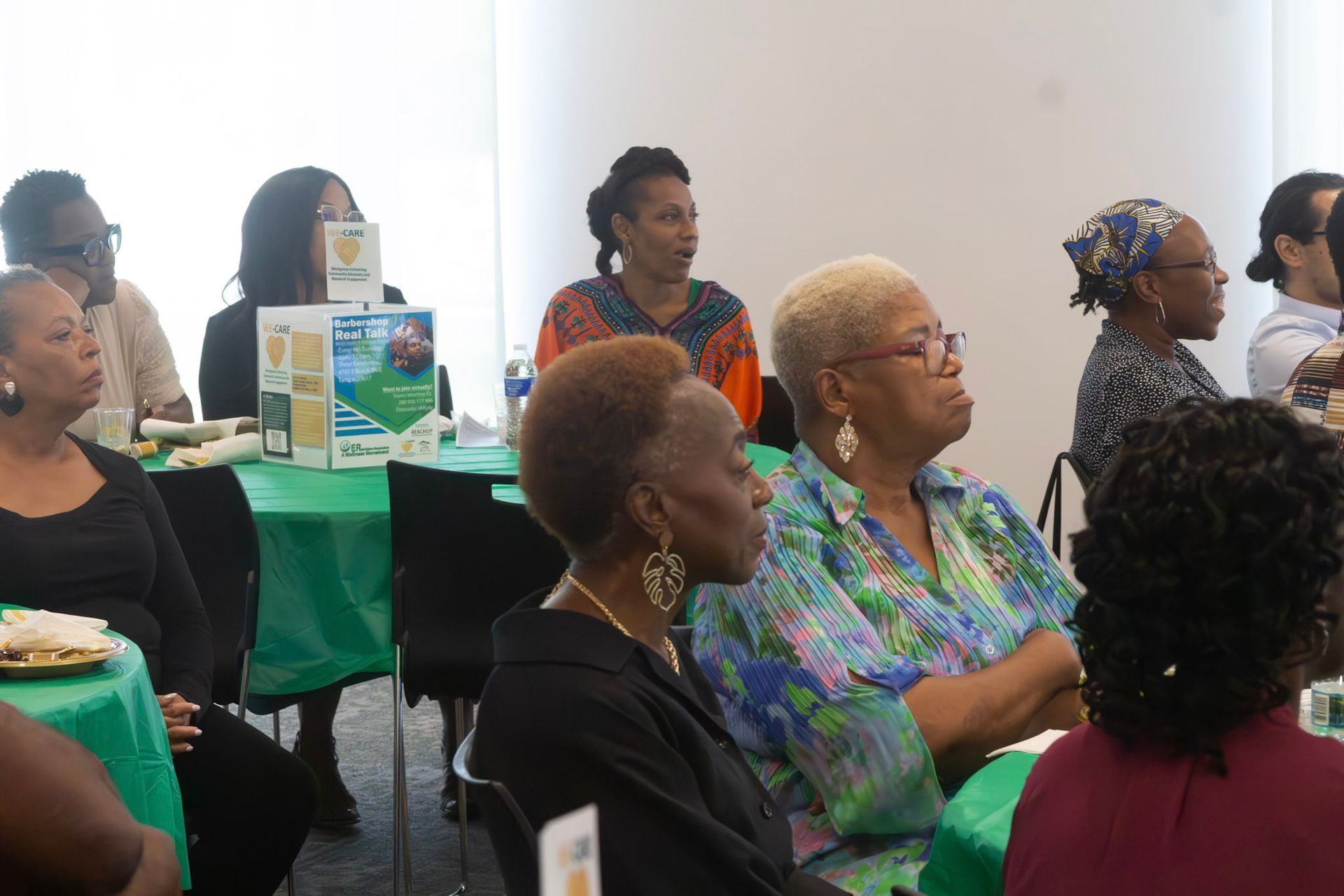 People seated at tables listening to a presentation in a room. Green table covers, sunlight.