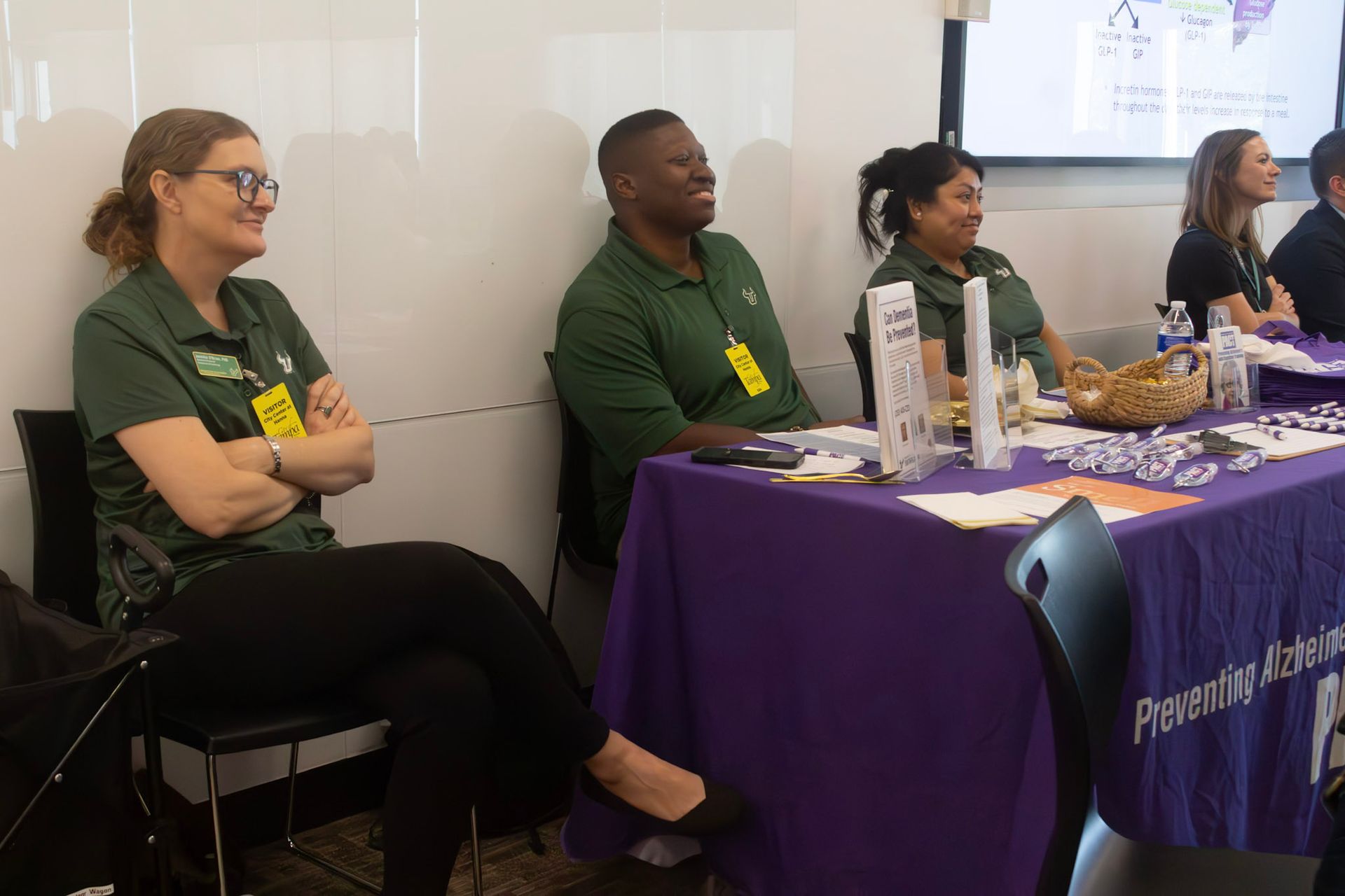 People at a table with purple tablecloth, presumably an event. Three wear green shirts. One with arms crossed.