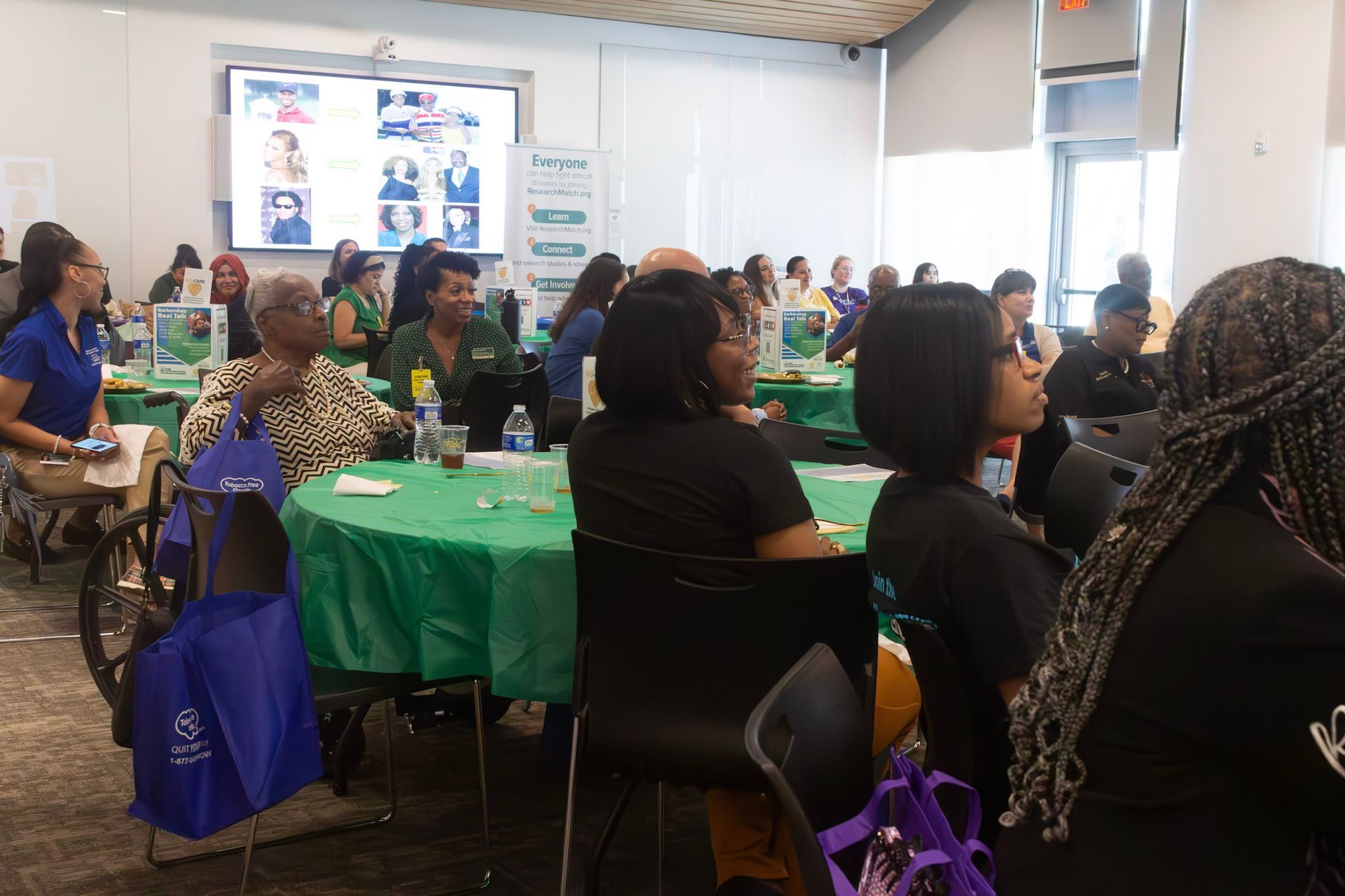 Attendees listen at an event with round tables covered in green. A screen displays photos.