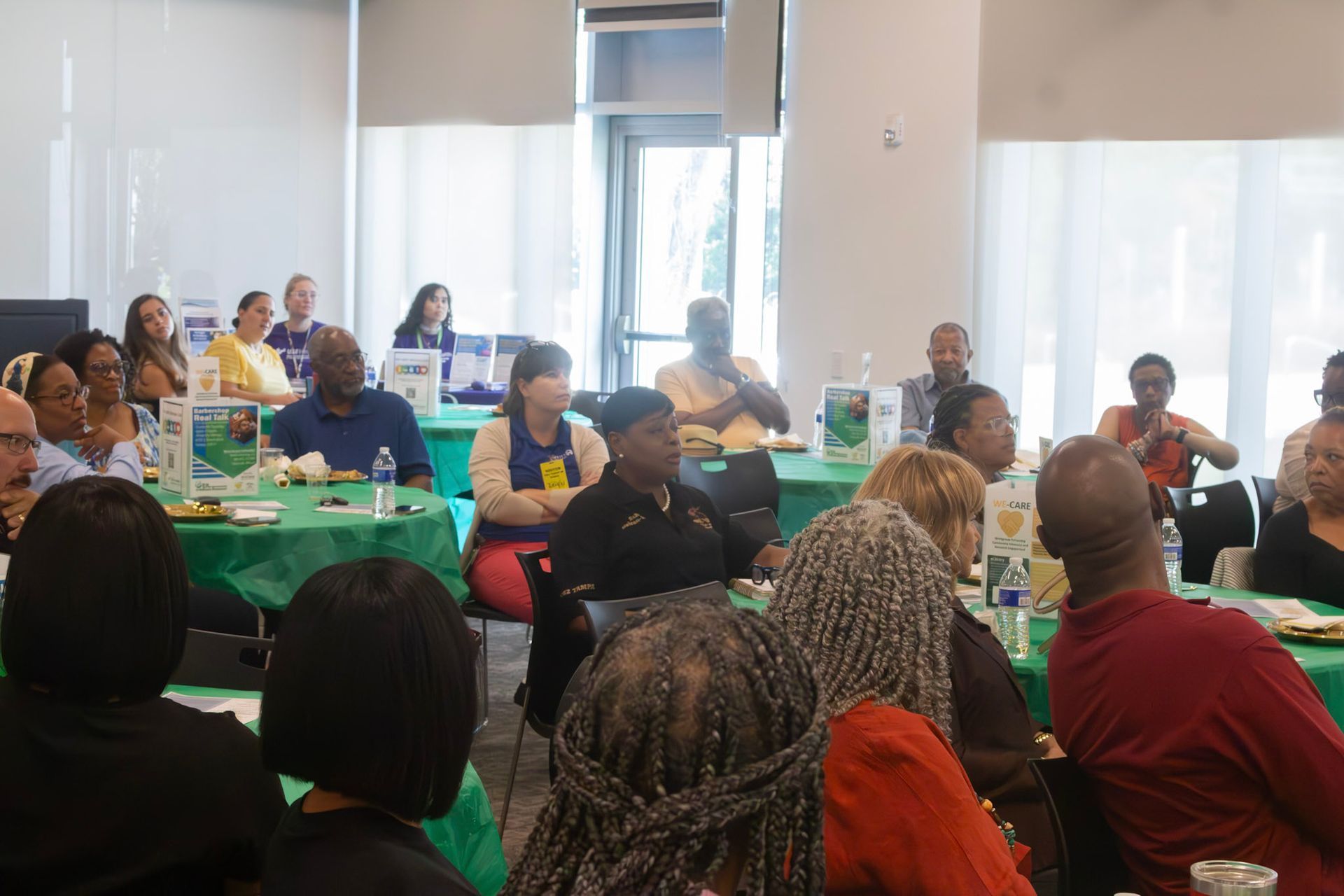 People seated at tables in a conference room with green tablecloths, attending a meeting.