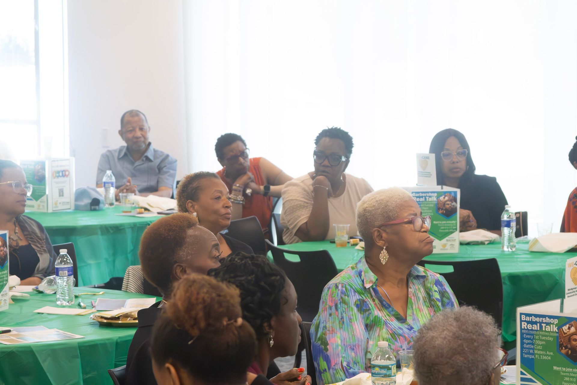 People seated at green tables in a well-lit room, possibly a meeting or presentation.