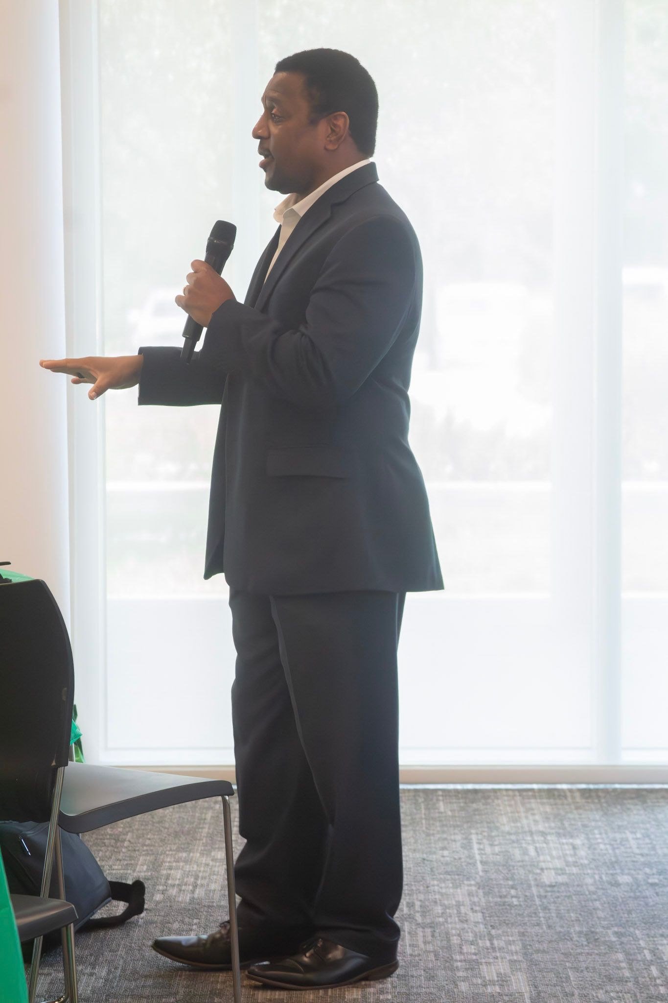 Man in a suit speaks into a microphone, gesturing with his left hand near a window.