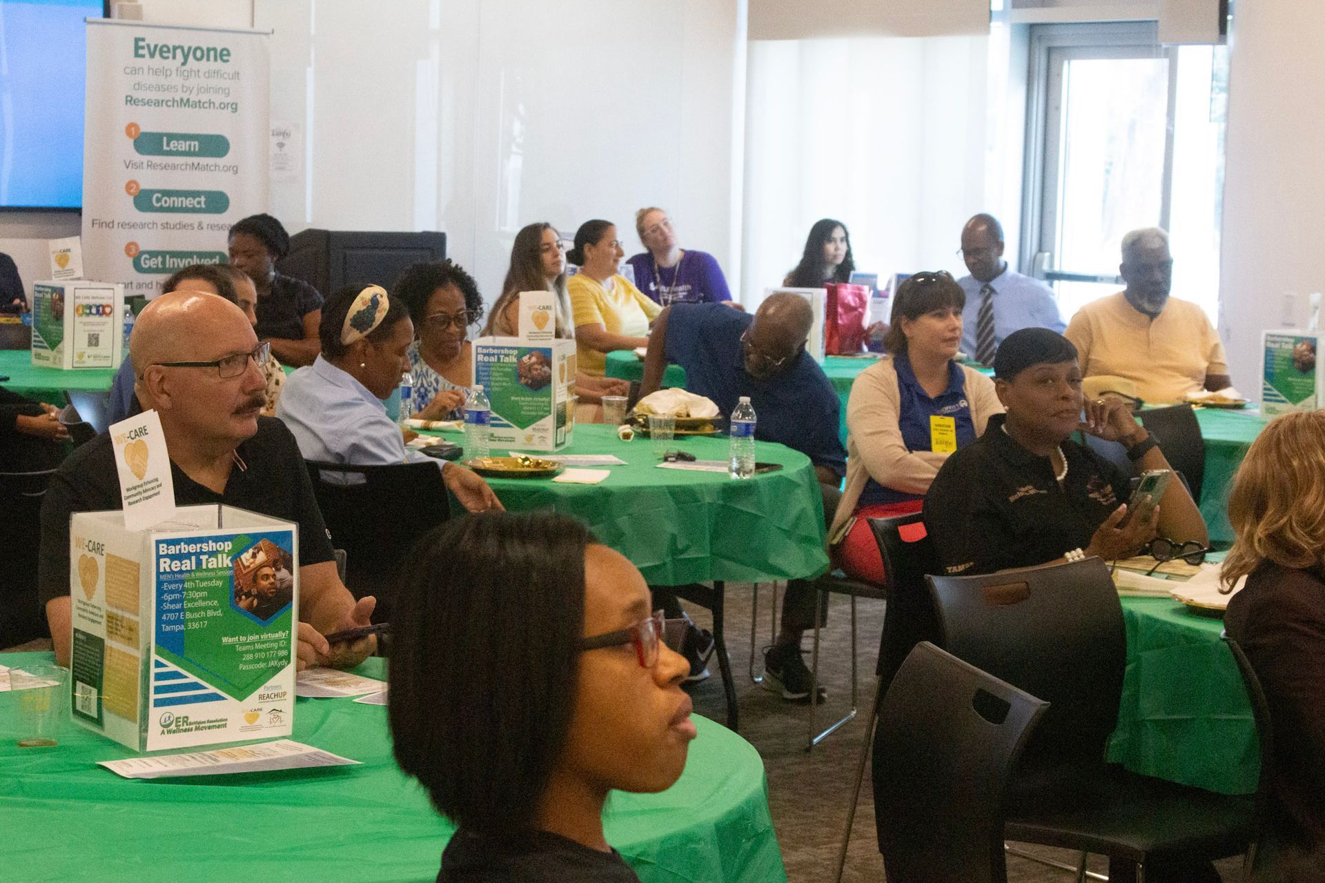 People seated at round green tables, attending a gathering, some looking forward, a banner in the background.
