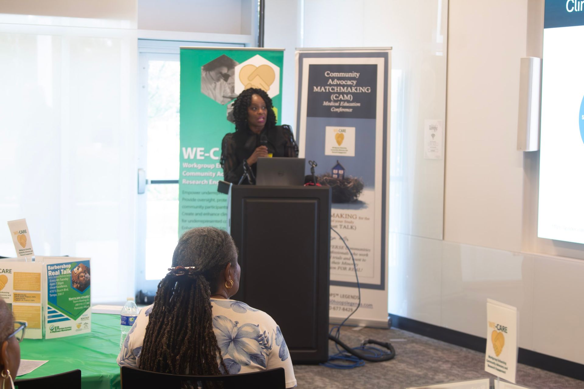 Woman presenting at a podium. Two attendees in foreground, promotional banners behind her, screen to the right.