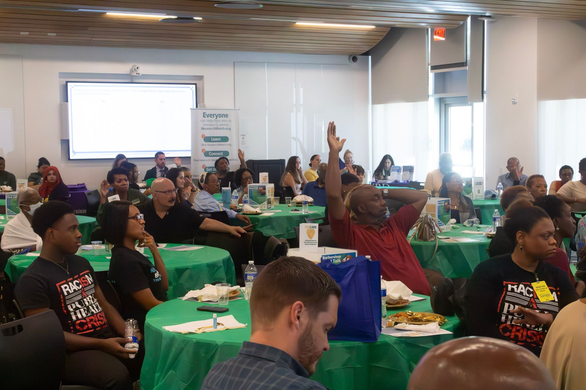 Audience at a conference raises hands, seated at green-covered tables, in a bright, modern room.