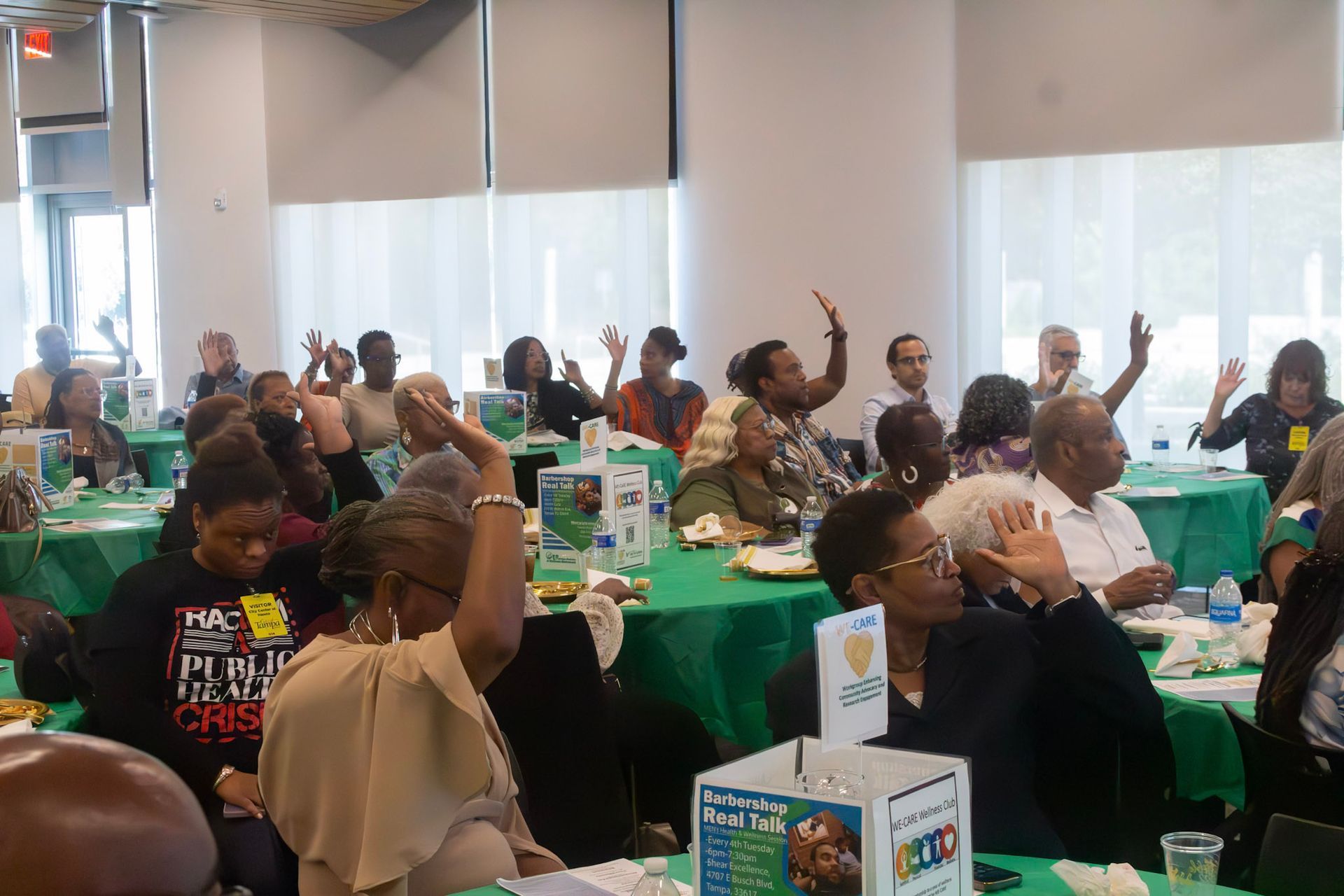People raising hands at tables covered in green cloths in a meeting.