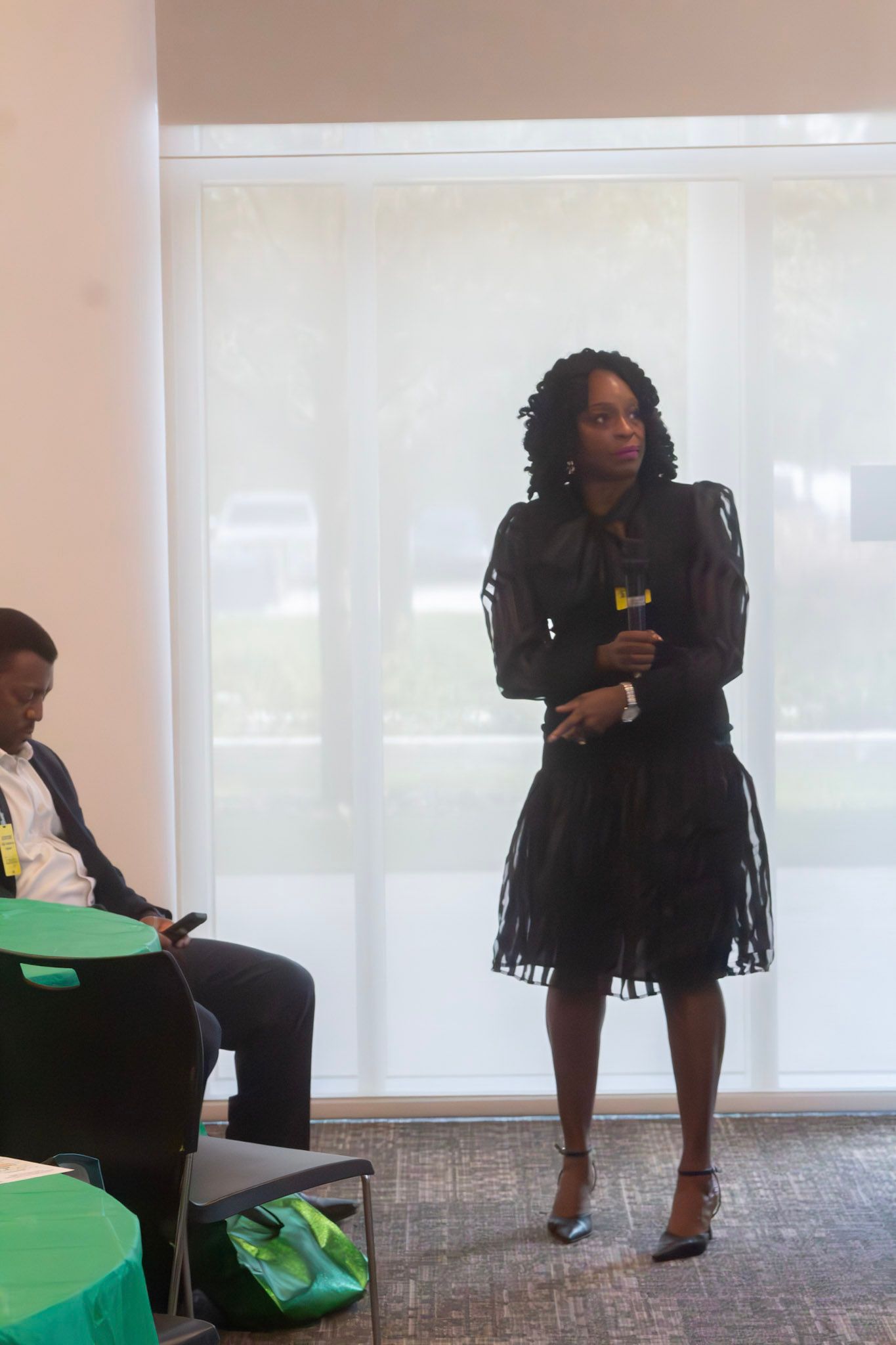 Woman in black dress stands and looks right. Man sits to the left. Bright room.
