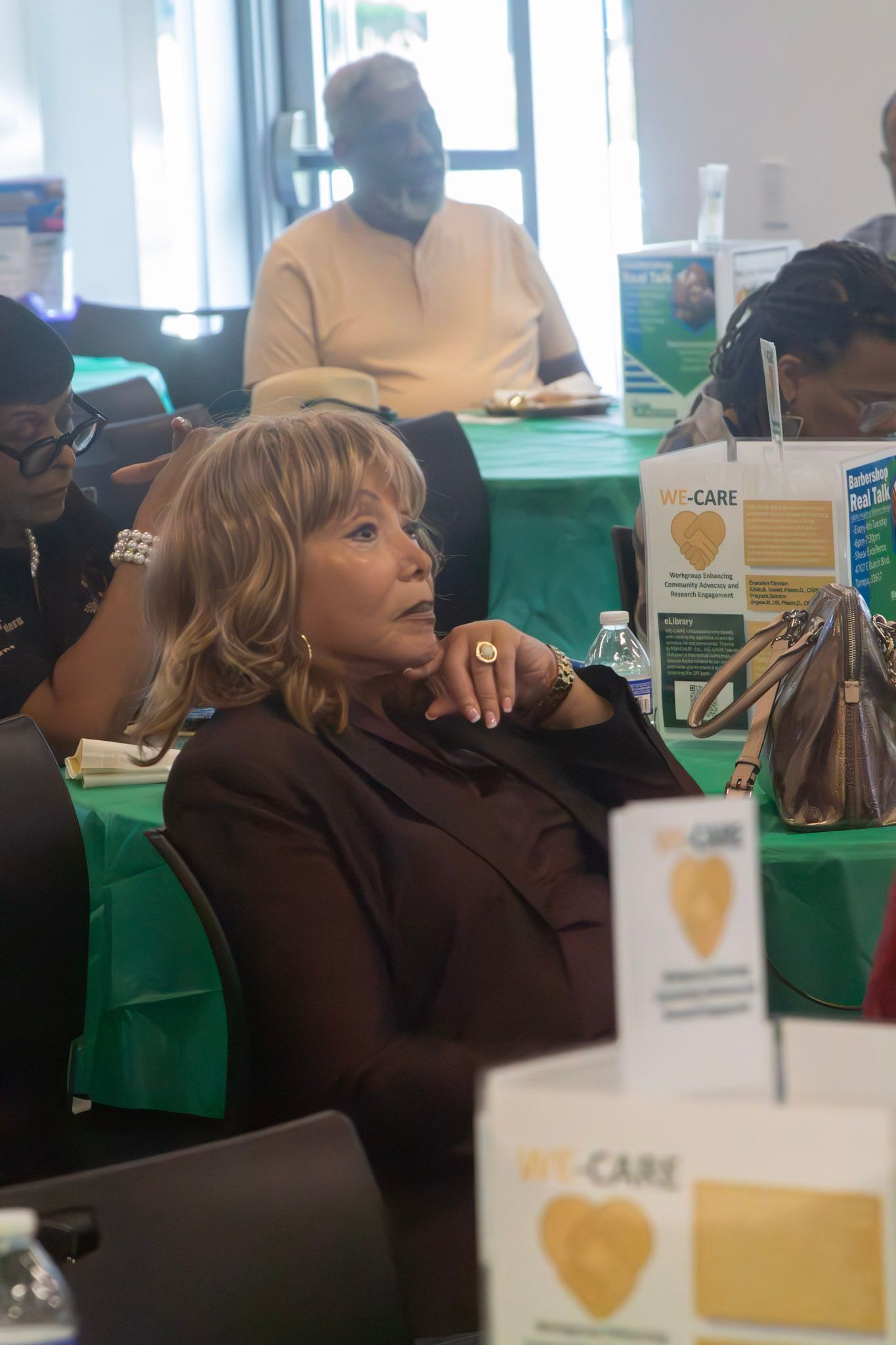 Woman in brown blazer at an event, seated, looking to the side. Other people and tables are in the background.