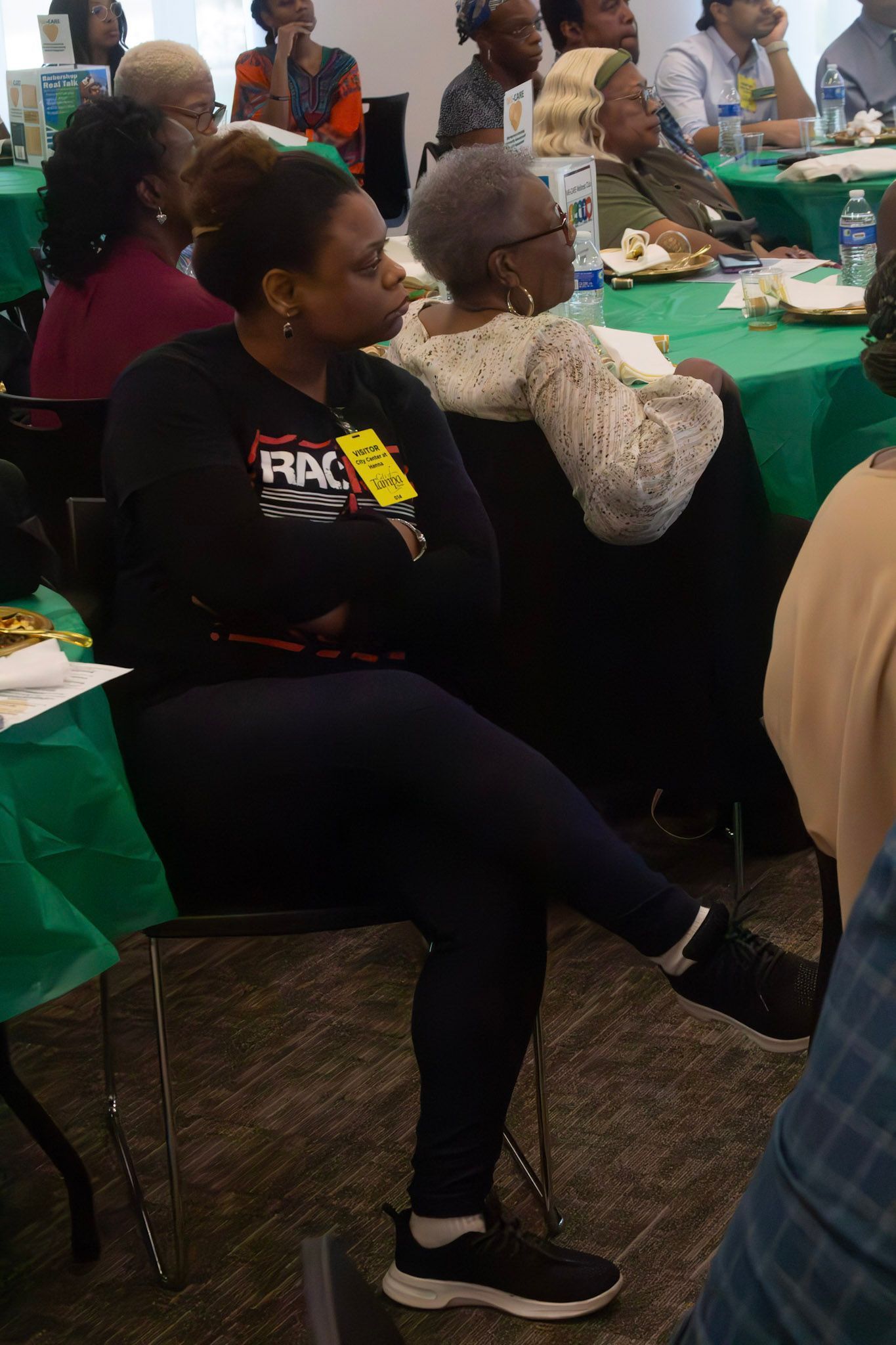 Woman in black shirt and leggings sits at a table, arms crossed, observing a meeting.