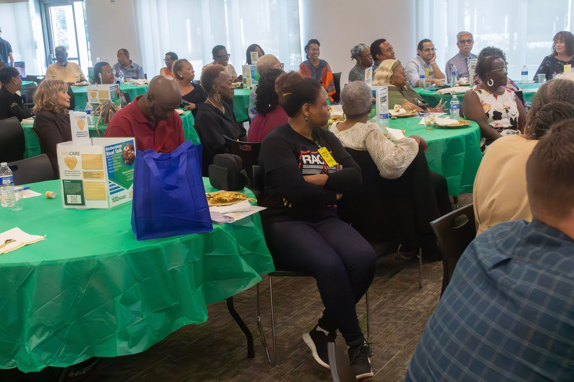 People seated at round tables in a brightly lit room; some are looking forward, others eating. Tables are covered in green.