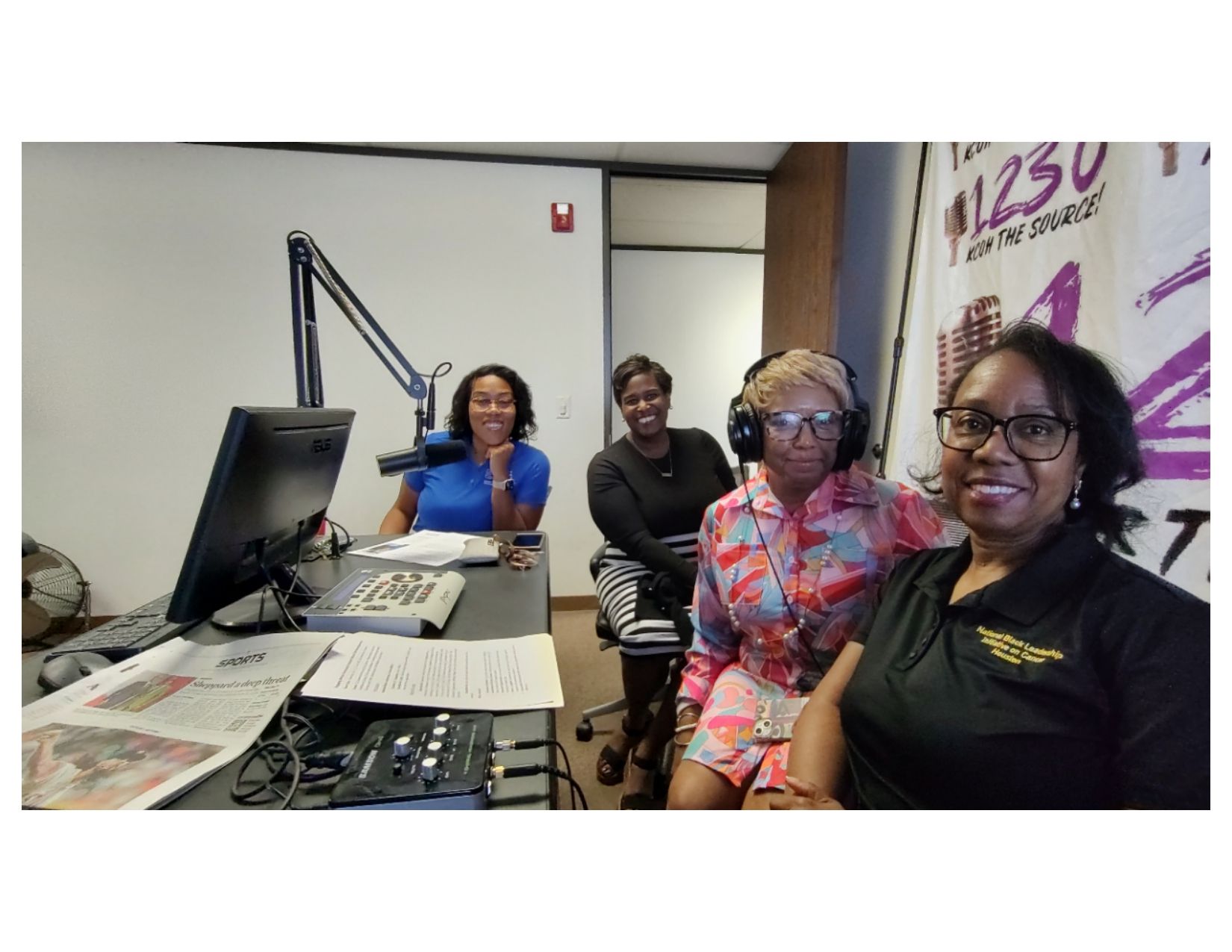 Four people in a radio studio. They sit at a desk with equipment. One wears headphones. A sign says 
