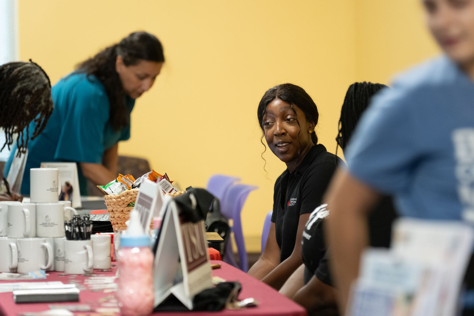 People at a table with mugs and literature; one woman smiles, others browse. Yellow wall in the background.