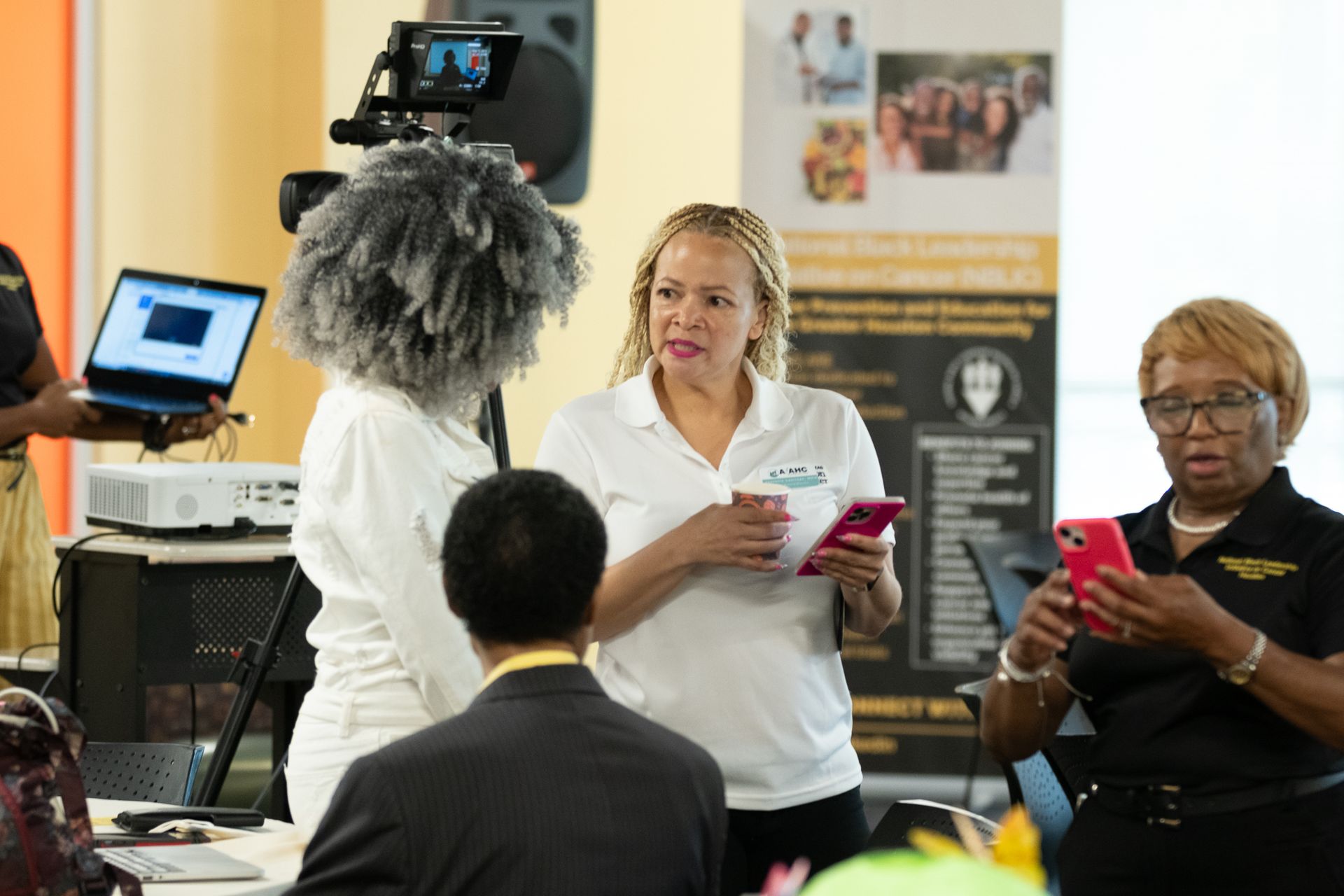 People at event: woman speaks, holds phone. Others with equipment, banner in background.