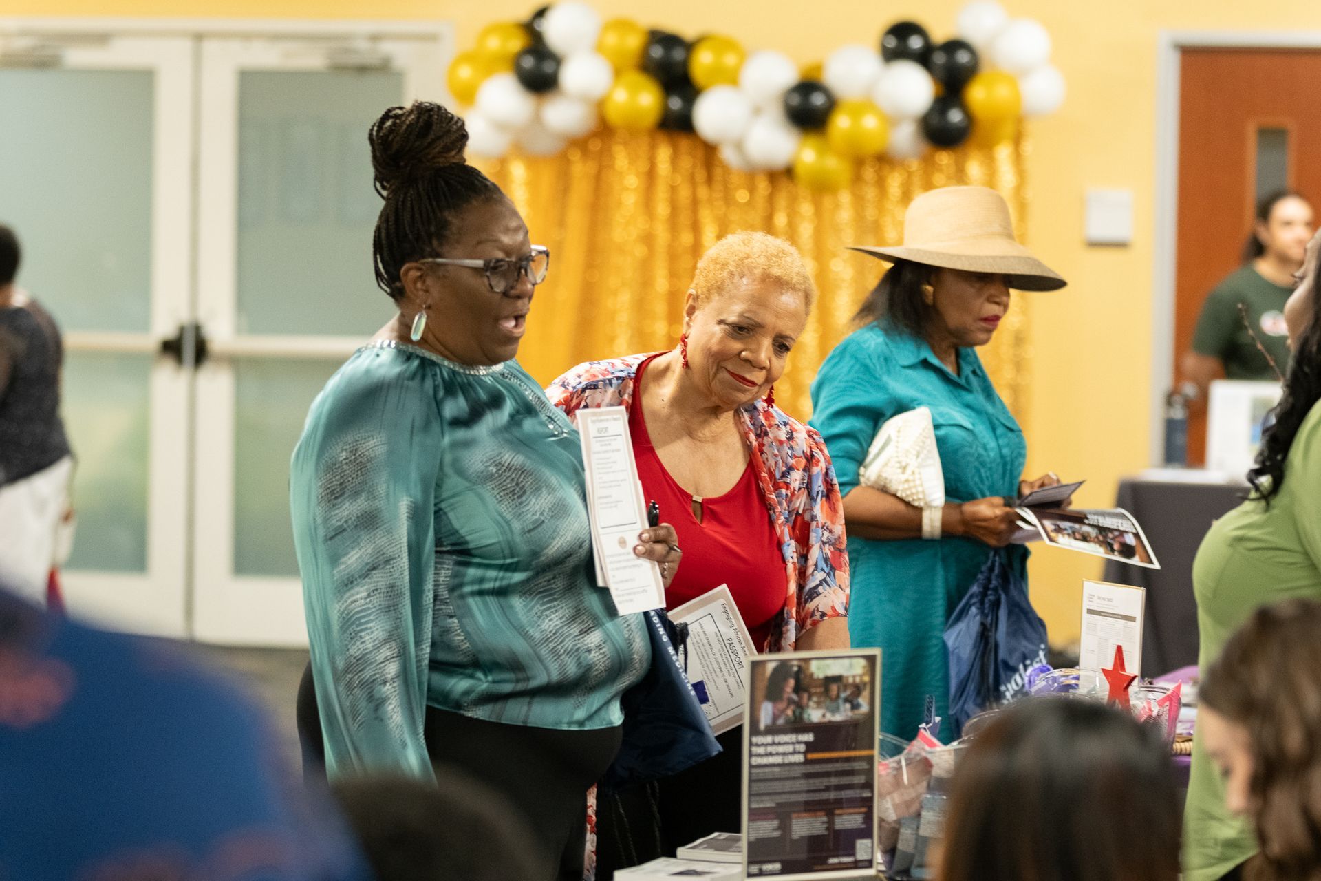 Three women look at materials at a table at an event. Balloons and a gold backdrop are behind them.
