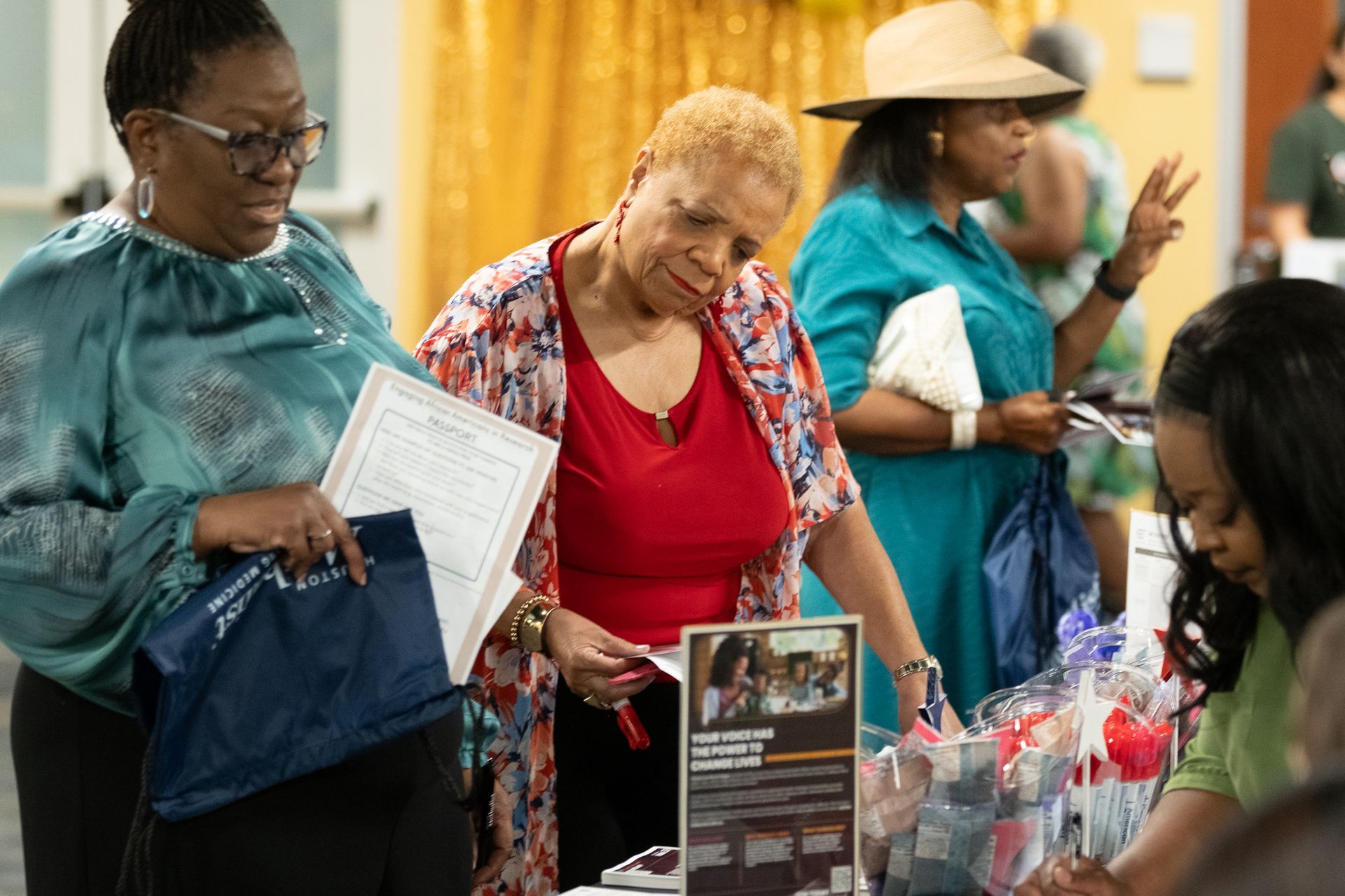 People looking at items on a table at an event. Woman in red shirt examines papers; others browse. Indoors, bright.