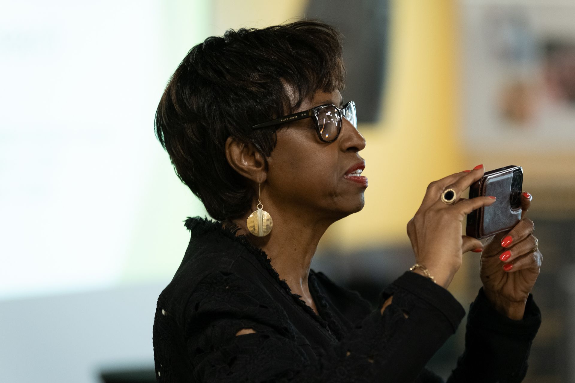 Woman with dark hair and glasses holding a phone, taking a photo. She is wearing a black jacket and large earrings.