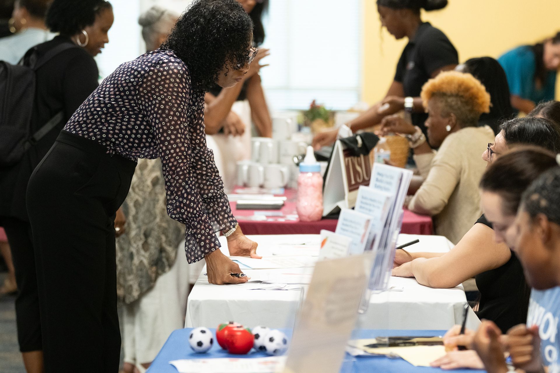 People at tables, likely an event. Woman in spotted shirt leans in. Others interact, reaching, writing.