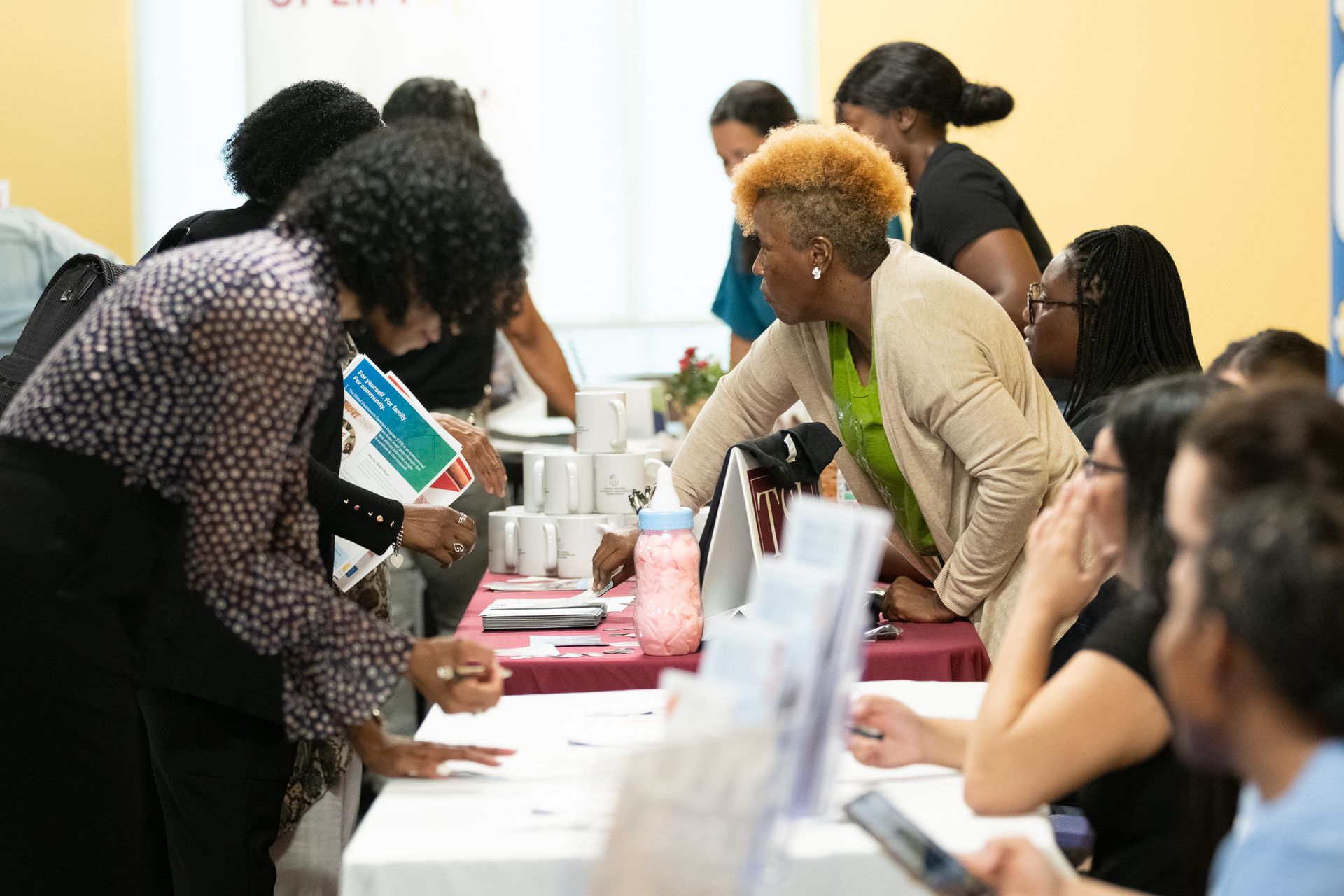 People at a table, some reaching for materials. Various mugs on table. Brightly lit room.
