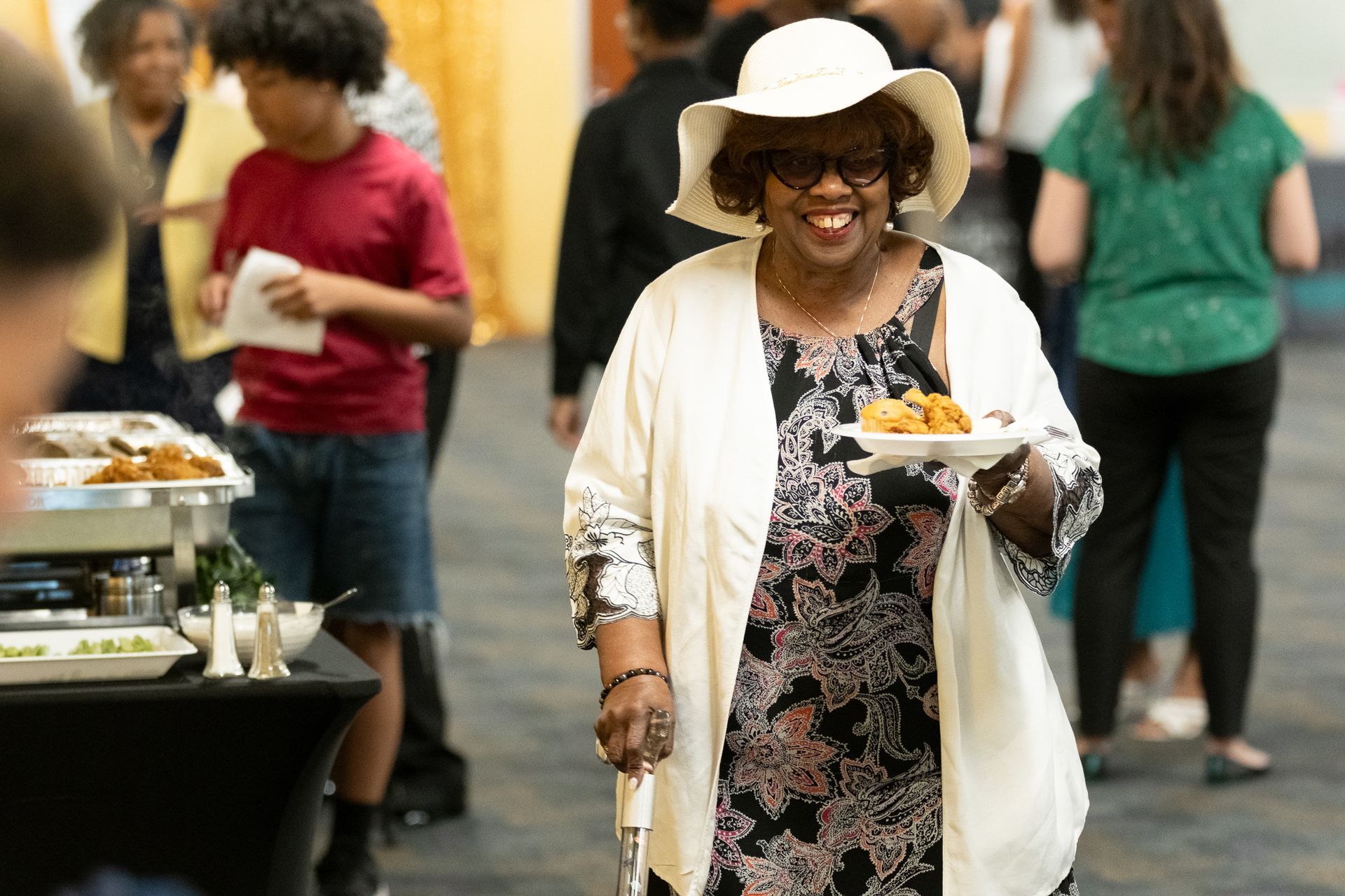 Woman smiling, holding plate of food, wearing a hat and cream-colored shawl at an event.