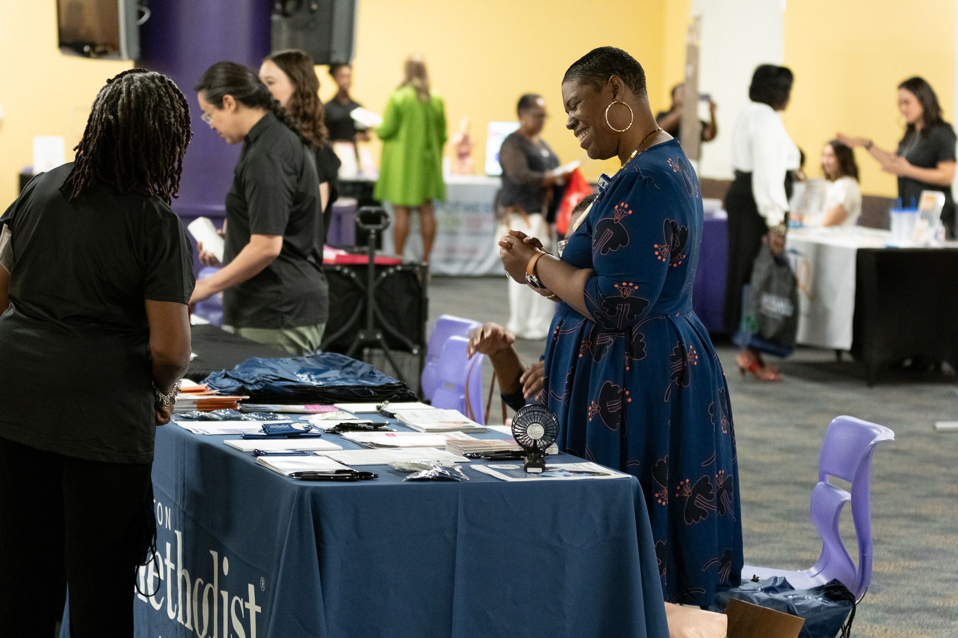 People at a booth, woman in blue dress looking at items. Event setting with tables and other people.