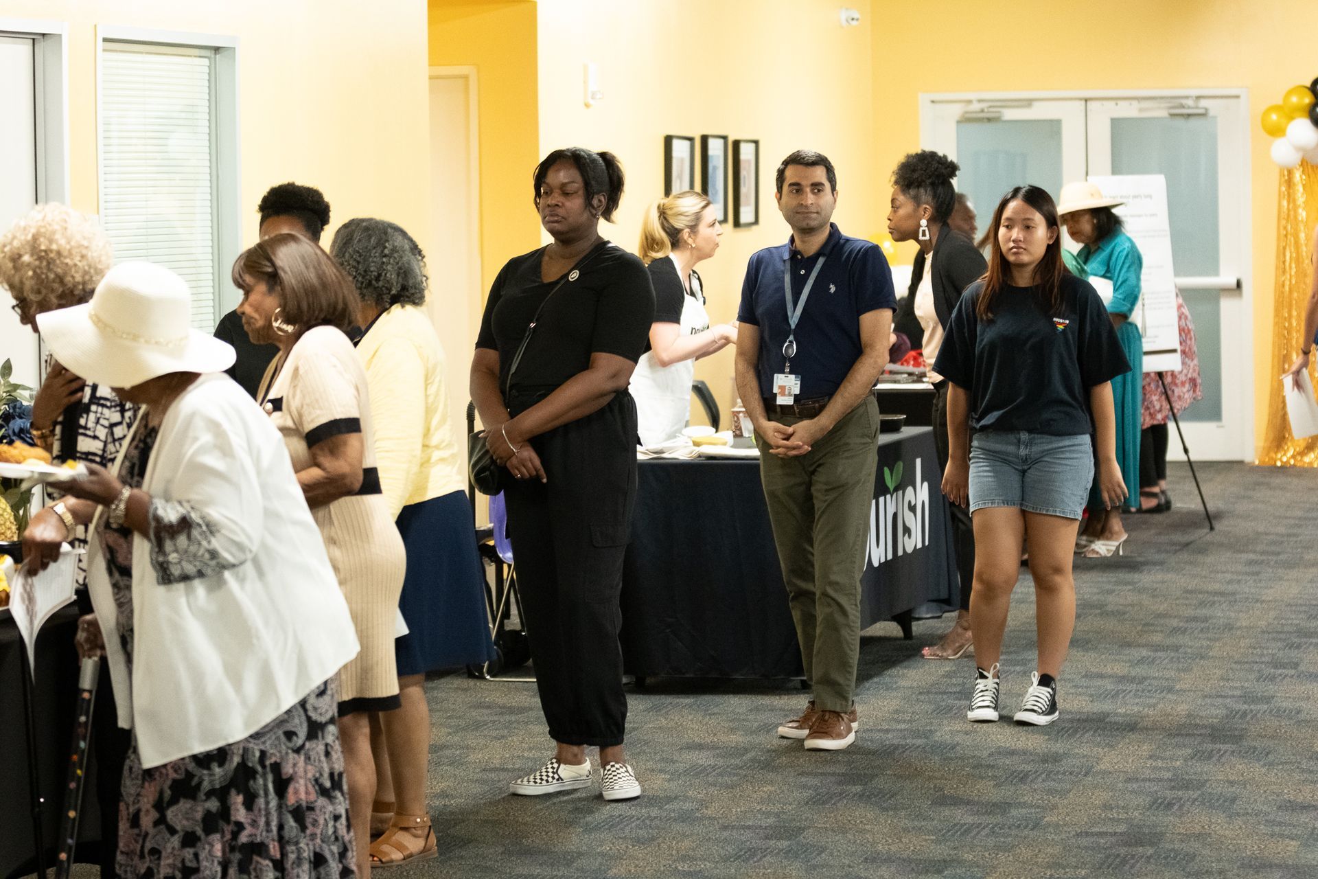 People at an event, standing and interacting. Food and decorations visible. Bright room.