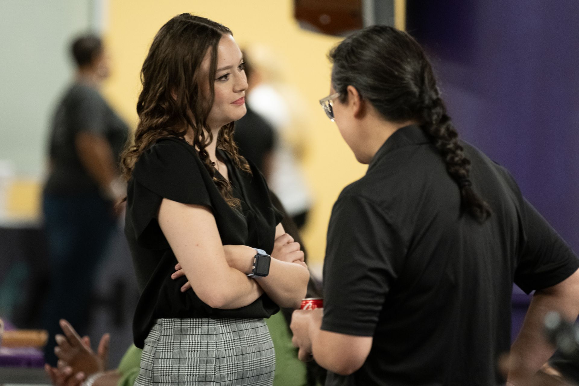 Woman in black shirt and plaid pants talks with a person with long braided hair and glasses