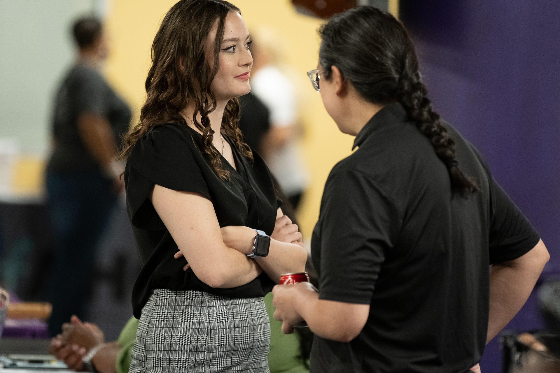 Woman with crossed arms talking to a person with braided hair in a room with a purple wall.