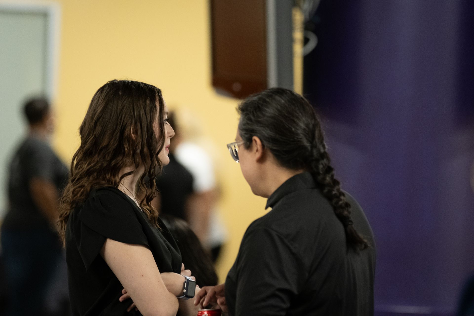 Two people in conversation; woman with wavy hair, man with long braid, in a colorful room.